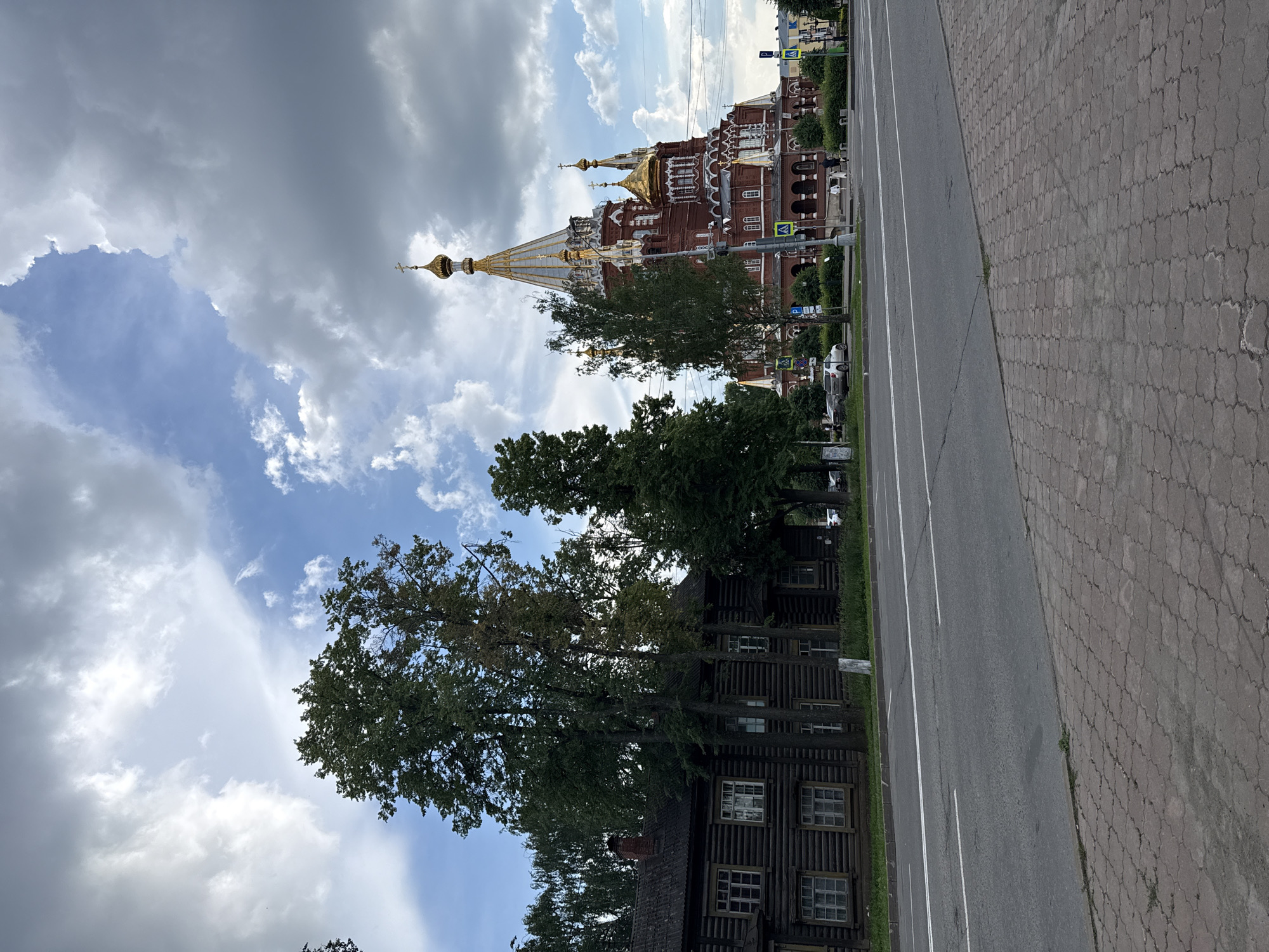 The image shows a street scene in Izhevsk, Russia, featuring a wooden building on the left and a red brick church with golden domes on the right. The foreground consists of a paved sidewalk and a road with white lane markings. Trees are present throughout the scene, with a few in the foreground and more in the background. The sky is partly cloudy with dramatic cloud formations. Cars are visible on the road and parked near the church. There are no prominent human figures engaged in specific actions. The overall impression is of a public space in a city, showcasing contrasting architectural styles. The wooden building appears to be a residential house, while the church is a religious structure. The street is well-maintained, and the environment suggests a normal day in the city.
