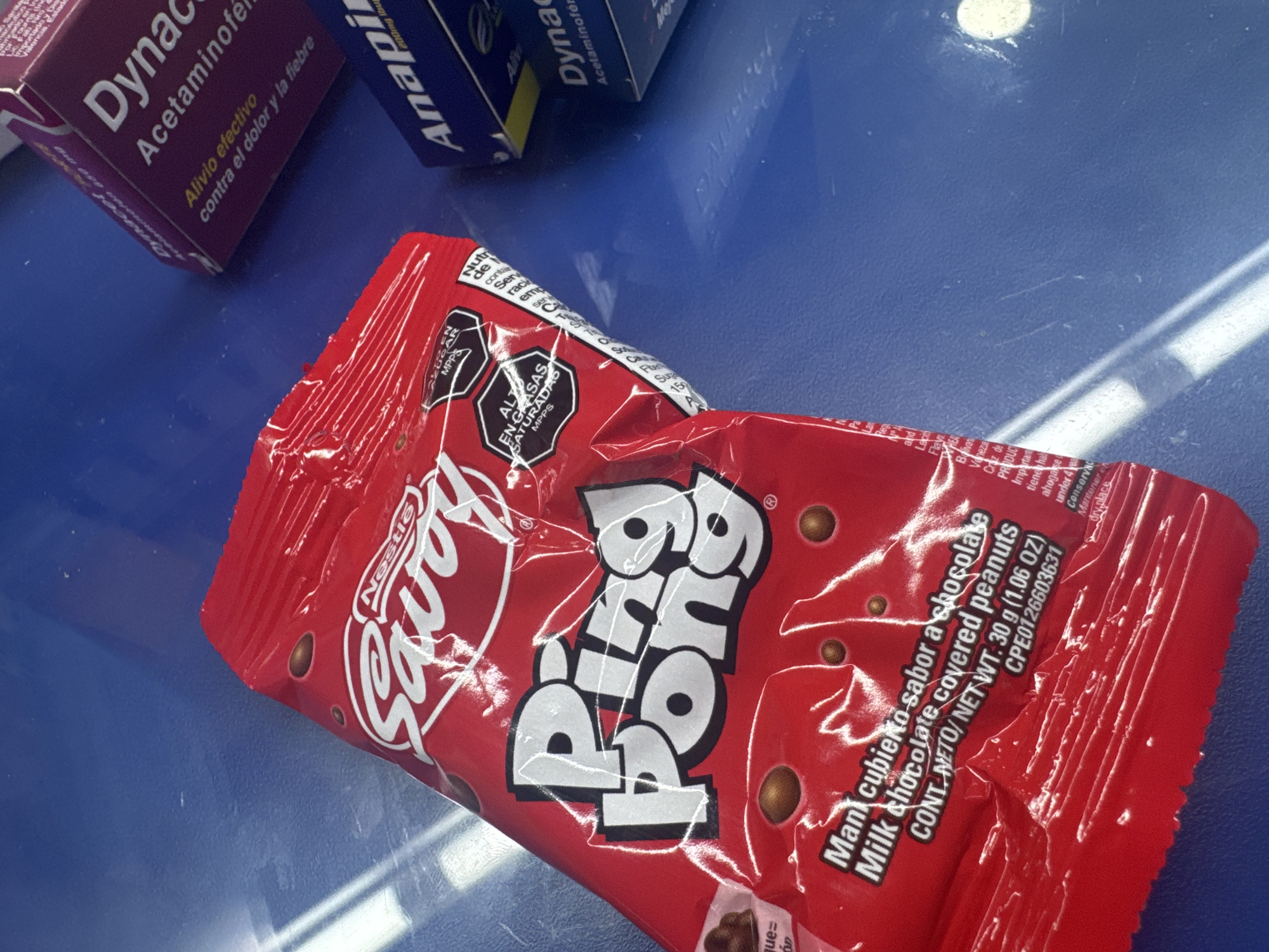 The image shows a red wrapper of a "Ping Pong" candy from Savoy Nestle on a blue surface. The package has Spanish text indicating it contains milk chocolate covered peanuts. To the left of the candy are two boxes of medication: "Dynac" and "Anapi," medications commonly used to reduce fever and pain. The details on the wrapper indicate the product is manufactured in Parroquia Ocumare, Venezuela. There is no information regarding people, subjects, or actions.