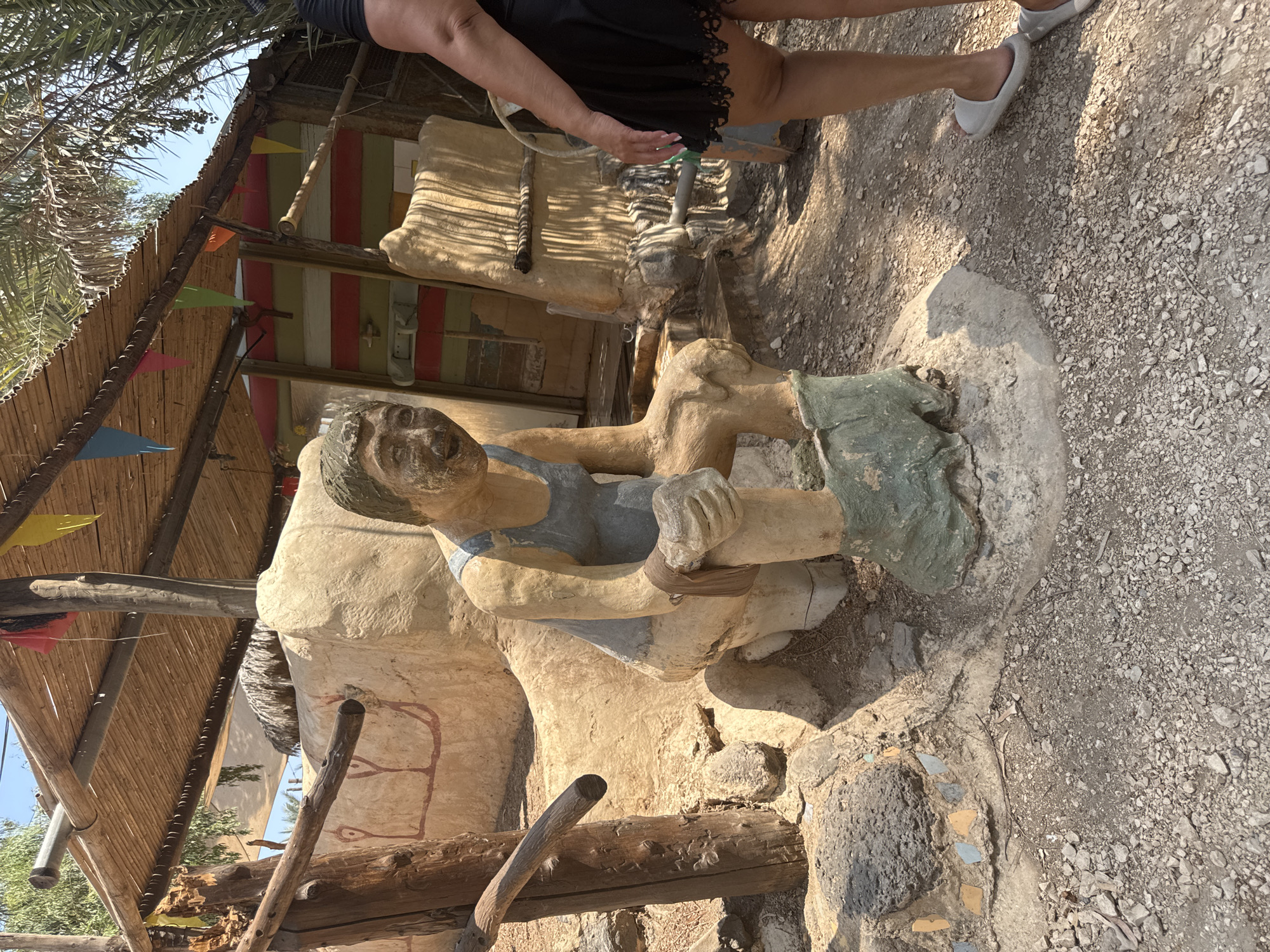 The image depicts an outdoor scene with a statue of a person sitting on a toilet. The statue is made of a tan material and is wearing a blue tank top and green pants that are partially pulled down. The statue has its hands on its knees, and its facial expression appears to be one of strain. The statue is placed in a sandy, rocky area with some vegetation in the background. A person's legs, wearing a black skirt and white slippers, are visible on the right side of the image, suggesting a real person is observing or interacting with the statue. The overall environment suggests a rural or potentially tourist-oriented location, possibly an amusement park or a roadside attraction, given the whimsical nature of the statue. The background includes a wooden structure with a thatched roof, adorned with colorful triangular flags. The location is specified as Emek HaYarden Regional Council, Israel.