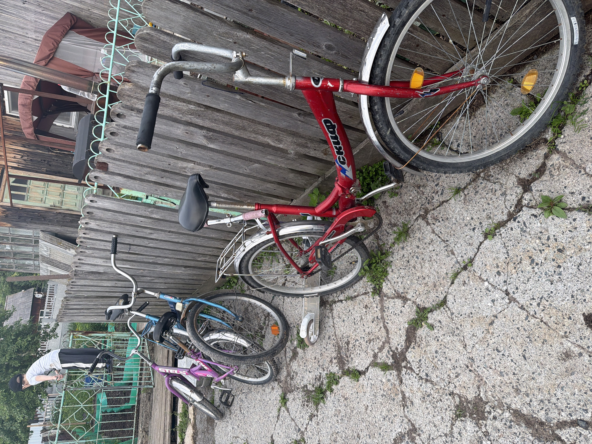 A young boy wearing a white t-shirt and black pants stands on a metal fence, looking down at something. In front of him, several bicycles are parked. A red bicycle is in the foreground, leaning against a wooden fence. To its left are two other bicycles, one blue and one pink, also leaning against the fence. A silver scooter lies on the ground between the red bicycle and the blue bicycle. The ground is paved with cracked concrete and patches of grass. The wooden fence behind the bicycles is weathered and appears to be made of vertical planks. In the background, there are buildings with wooden siding and a partially visible brown canopy. The scene appears to be an outdoor residential area, possibly a backyard or side yard.