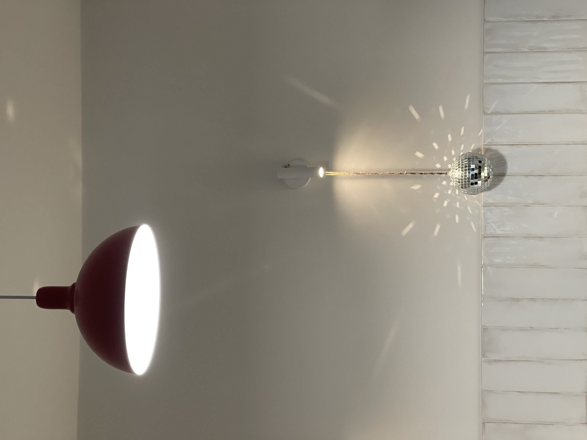 The image shows an interior with a red pendant lamp with a large, illuminated bulb hanging on the left. A white wall extends to the right, where a disco ball is mounted near a section of white, horizontally laid tile. The disco ball has light reflecting off its mirrored surfaces. A white wall-mounted fixture seems to power the disco ball. The photograph appears to be taken indoors. There is no information to locate the image in the city of Izhevsk, Russia.