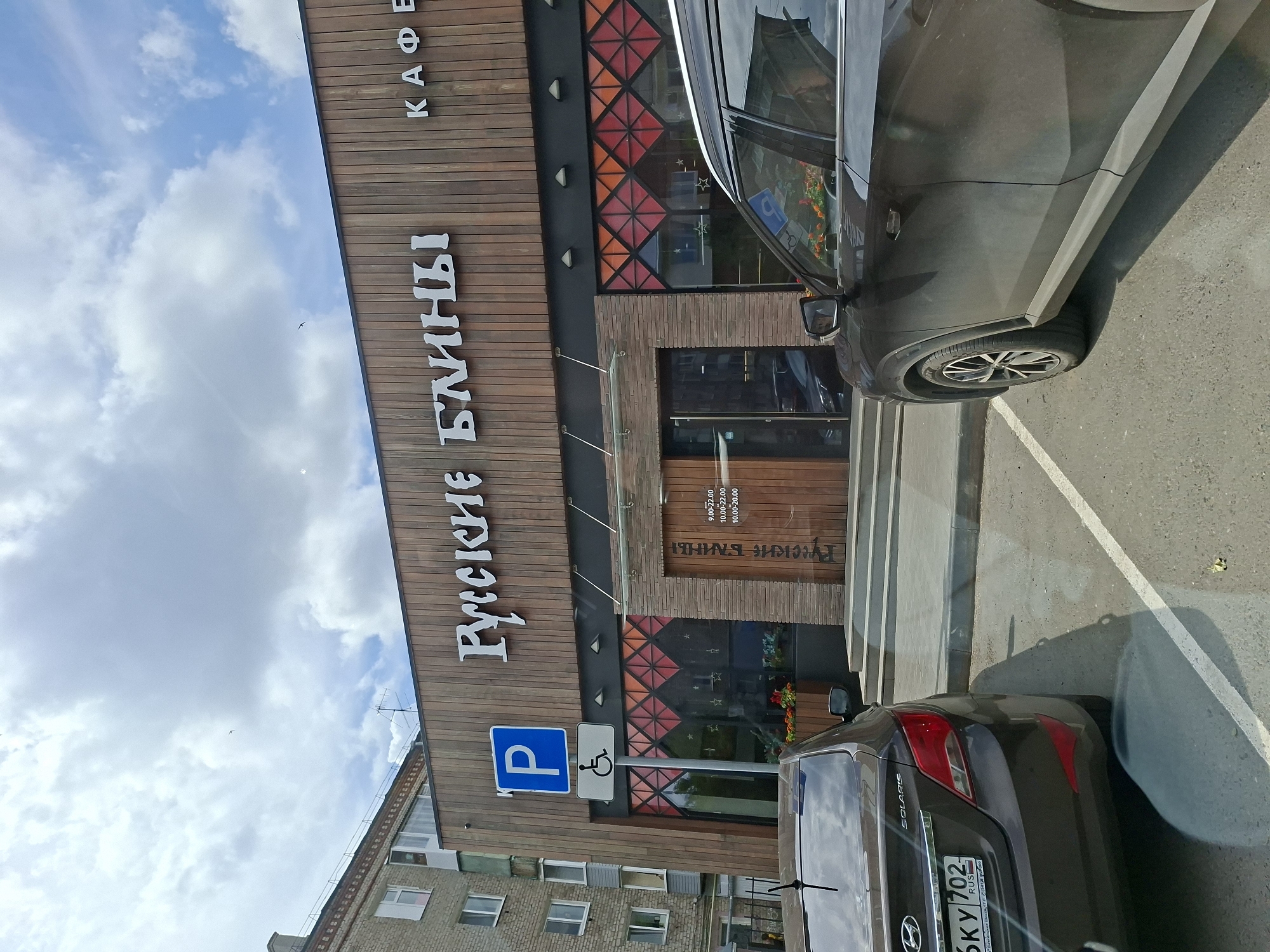 The image shows the exterior of a cafe named "Русские Блины" (Russian Pancakes) in Izhevsk, Russia. The cafe has a modern facade with wooden paneling and decorative stained-glass windows. A parking sign and a disabled parking sign are visible to the left of the entrance. Two cars are parked in front of the cafe. The sky is cloudy with patches of blue. The building appears to be in a commercial or residential area of the city. The overall impression is a casual dining establishment.