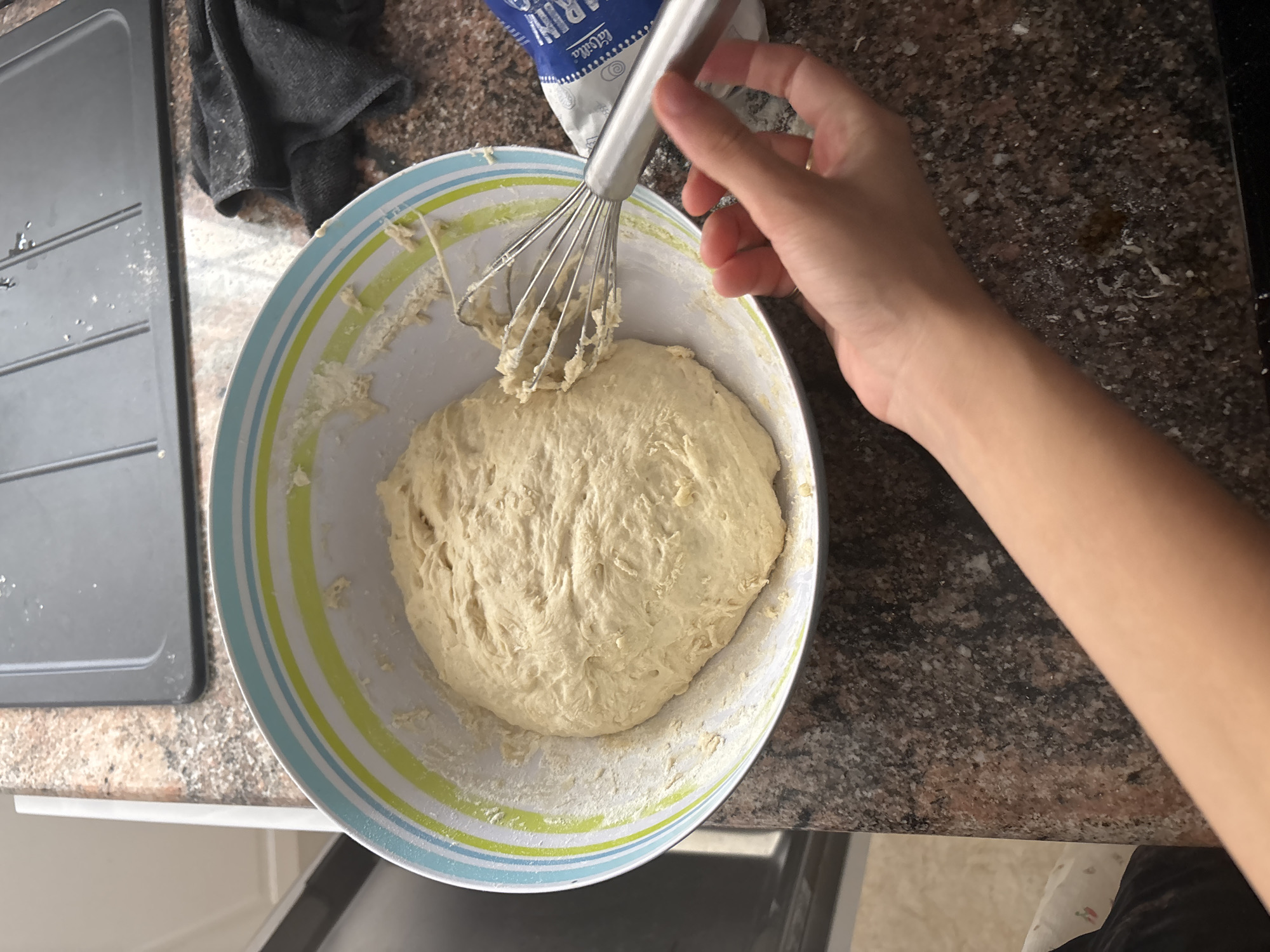 A person's hand and forearm are visible in the lower right corner of the frame, reaching towards a metal whisk that is partially submerged in a bowl of dough. The bowl is large and white with blue and green stripes around its rim. The dough appears to be light-colored and slightly lumpy. In the background, a dark grey countertop is visible, as well as a stainless steel sink and a dark cloth. A blue and white packet is also visible on the countertop to the right of the bowl. The overall scene appears to be a kitchen environment.