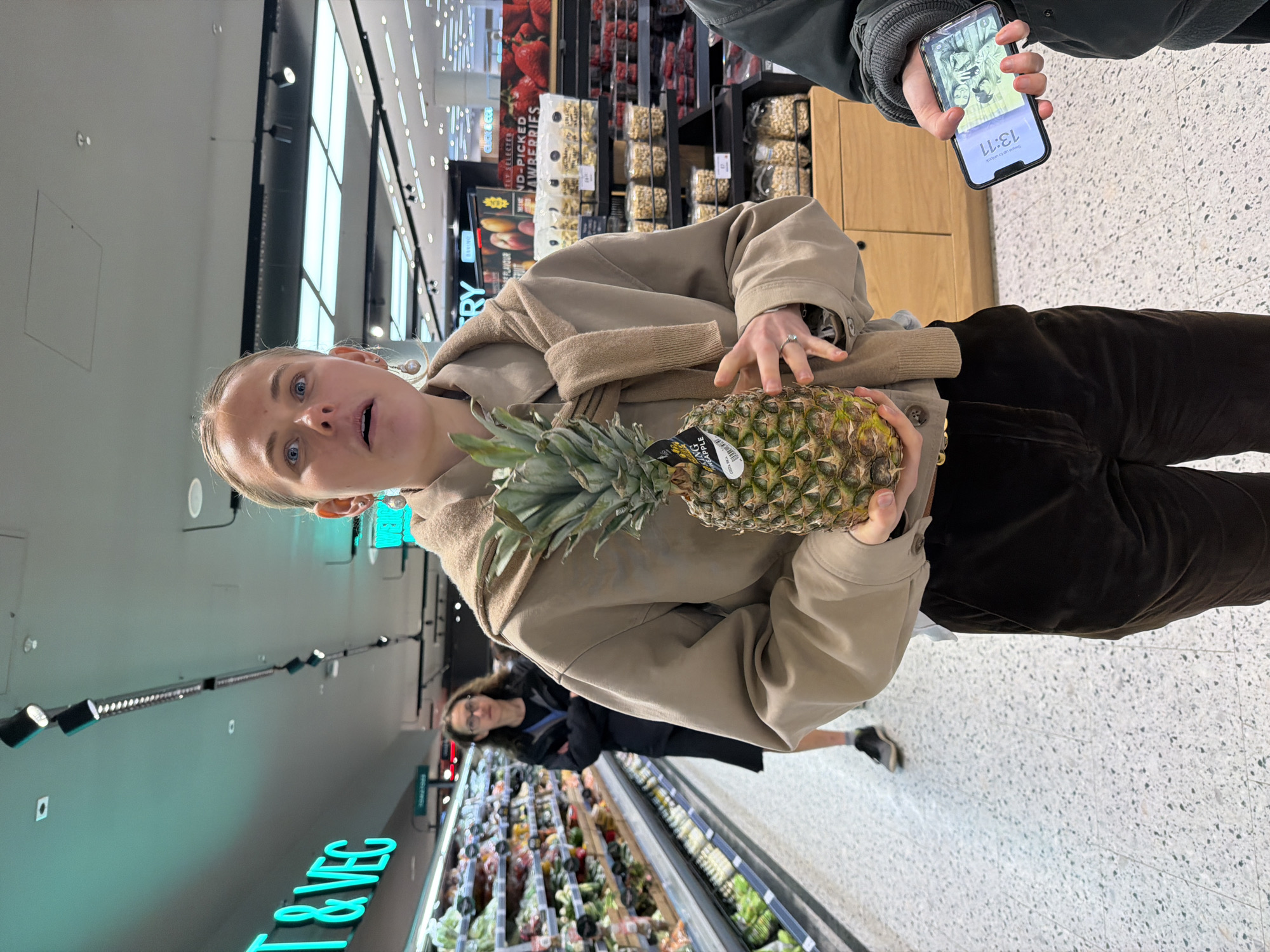 A tilted, medium-shot image captures a young, fair-skinned woman in a supermarket, holding a pineapple. She has blonde hair pulled back from her face and is wearing a beige jacket over a sweater. Her mouth is open slightly, and her blue eyes are wide. In the background, a person is holding a smartphone displaying a screen with an image and the time 13:11. The smartphone holder is wearing a dark-colored jacket. To the left, supermarket signage reads "T & VEC" in bright turquoise, with shelves of produce visible below. Another person is seen walking down the aisle, appearing to be shopping. The location appears to be a grocery store. Signage for "strawberries" can be seen in the background.