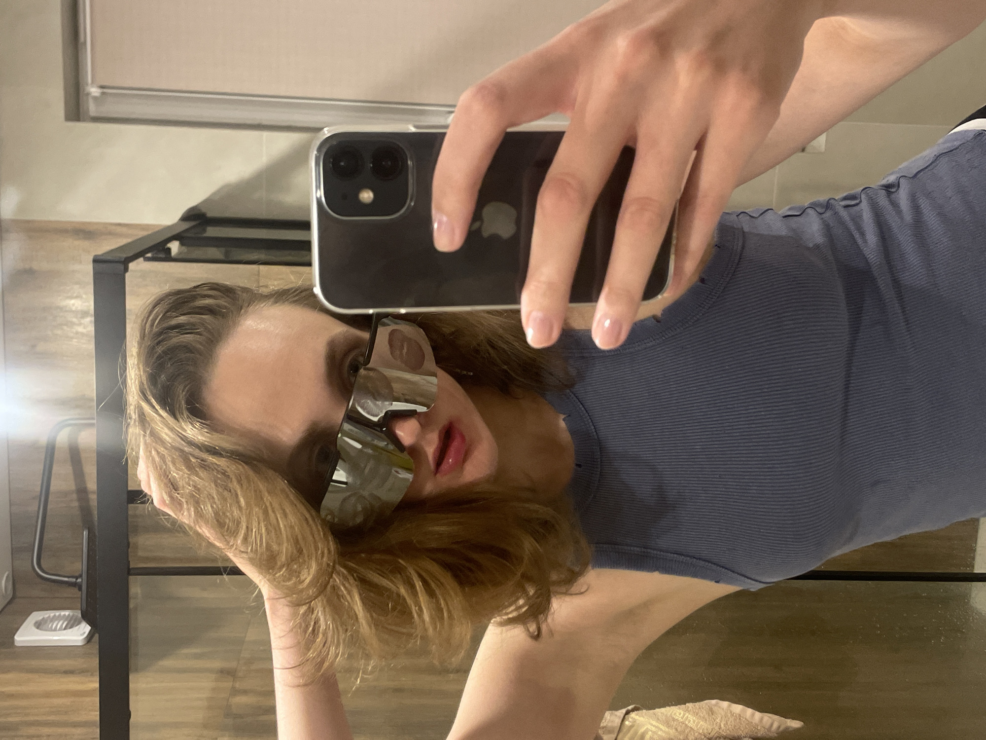 A person is taking a selfie in a bathroom, with the camera held up close to their face. The individual is wearing sunglasses and a blue tank top. Their hair is blonde and wavy. They are standing in front of a bathroom counter and mirror. A window or light fixture is visible in the background. The location is Unknown, Unknown.