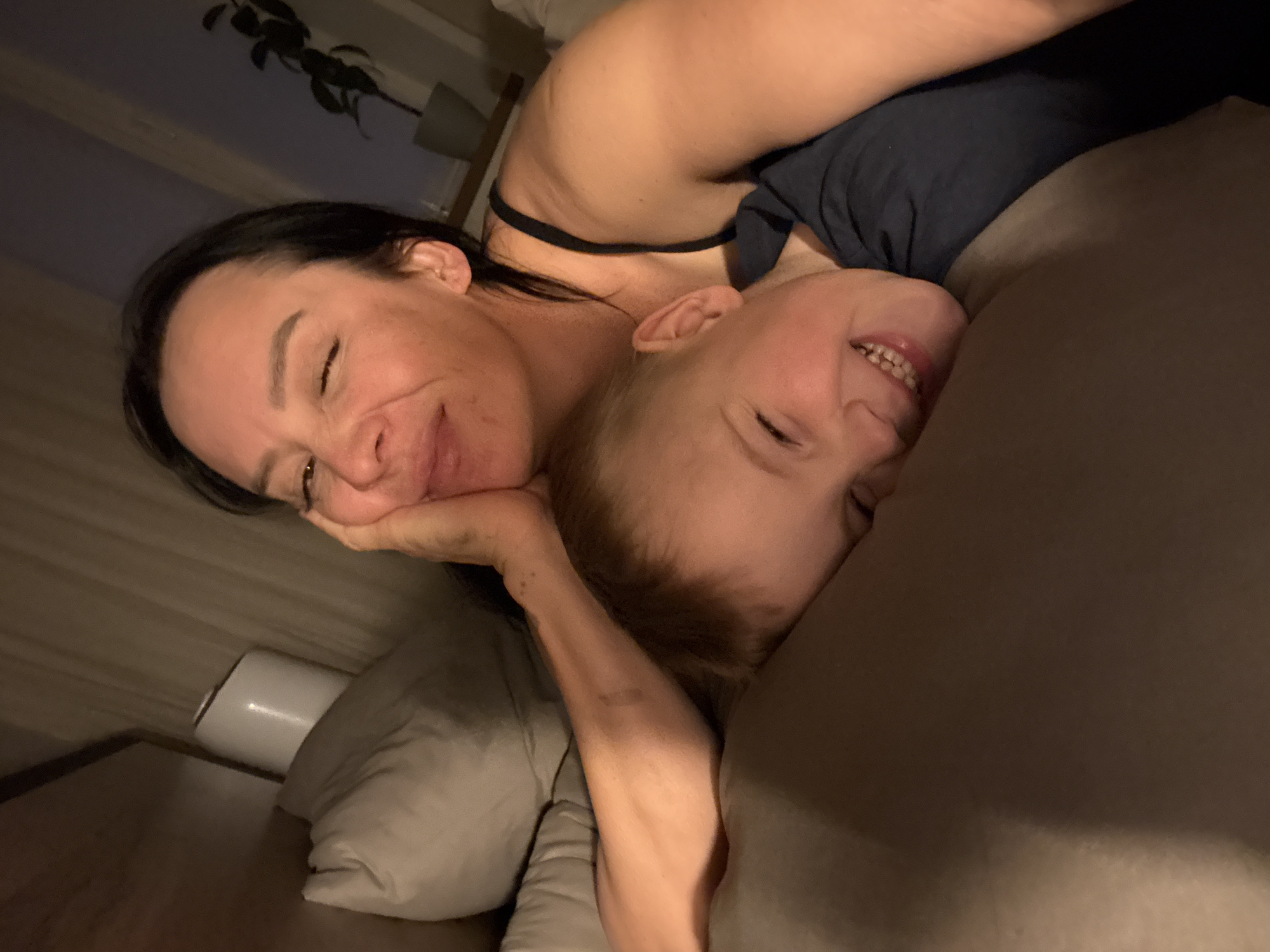 An indoor, eye-level shot captures a woman and a young child lying close together, both smiling widely, suggesting a moment of affection and joy. The woman, with dark hair, rests her head on her hand, looking towards the viewer with a soft smile. The child, with light hair, is snuggled against her, displaying a bright, toothy grin. They appear to be on a bed, surrounded by pillows and blankets. The dim, warm lighting indicates it is likely evening or nighttime. In the background, parts of a room are visible, including a dark potted plant and what appears to be a bedside lamp or light fixture. The overall atmosphere is intimate and happy. No visible text is present. The provided location context is Chaikovsky, Russia.