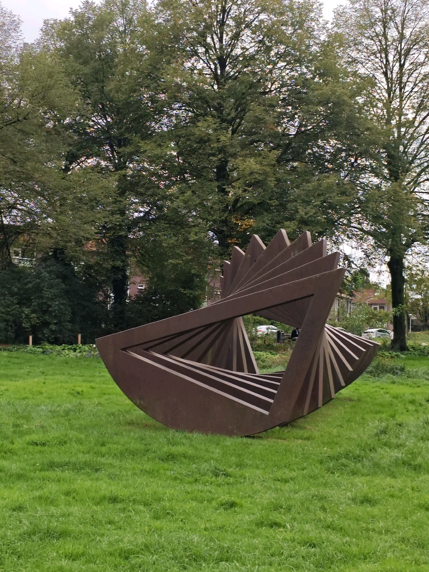 The image depicts an outdoor sculpture in a grassy area, likely a park or public space. The sculpture is made of a weathered, reddish-brown material, possibly metal, and has a complex, geometric form resembling a twisted helix or a series of fanned-out planes. It is positioned in the center of the frame, with lush green grass surrounding it. In the background, several large trees with full canopies dominate the scene. Behind the trees, houses with terracotta roofs and other buildings are visible, suggesting an urban or suburban setting. The sky is overcast, contributing to the muted lighting of the scene. There are no people visible in the image. The sculpture's design is abstract and industrial, contrasting with the natural elements of the grass and trees. The overall impression is one of public art integrated into a green urban environment. This sculpture is located at the John F. Kennedyplein in Eindhoven, Netherlands.