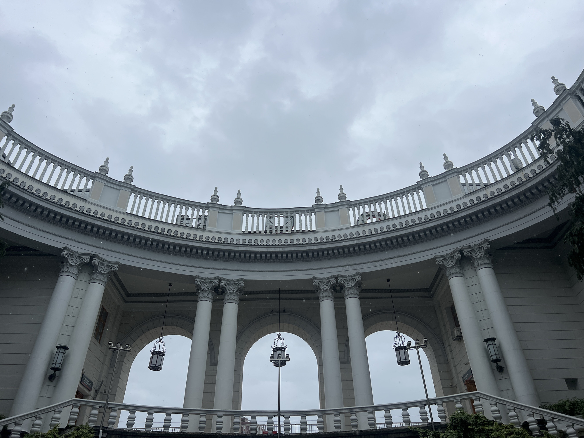 The image shows the colonnade of the Sochi railway station in Russia, taken from a low angle looking upwards. The architecture is classical, with white columns topped with ornate capitals. Above the columns is a balustrade with decorative finials. The sky is overcast and grey. Several hanging lanterns are suspended within the arches formed by the columns. A second level balustrade is visible, with vegetation peeking up from behind it. Fine snow or rain is falling. The image captures the symmetry and grandeur of the building's design.