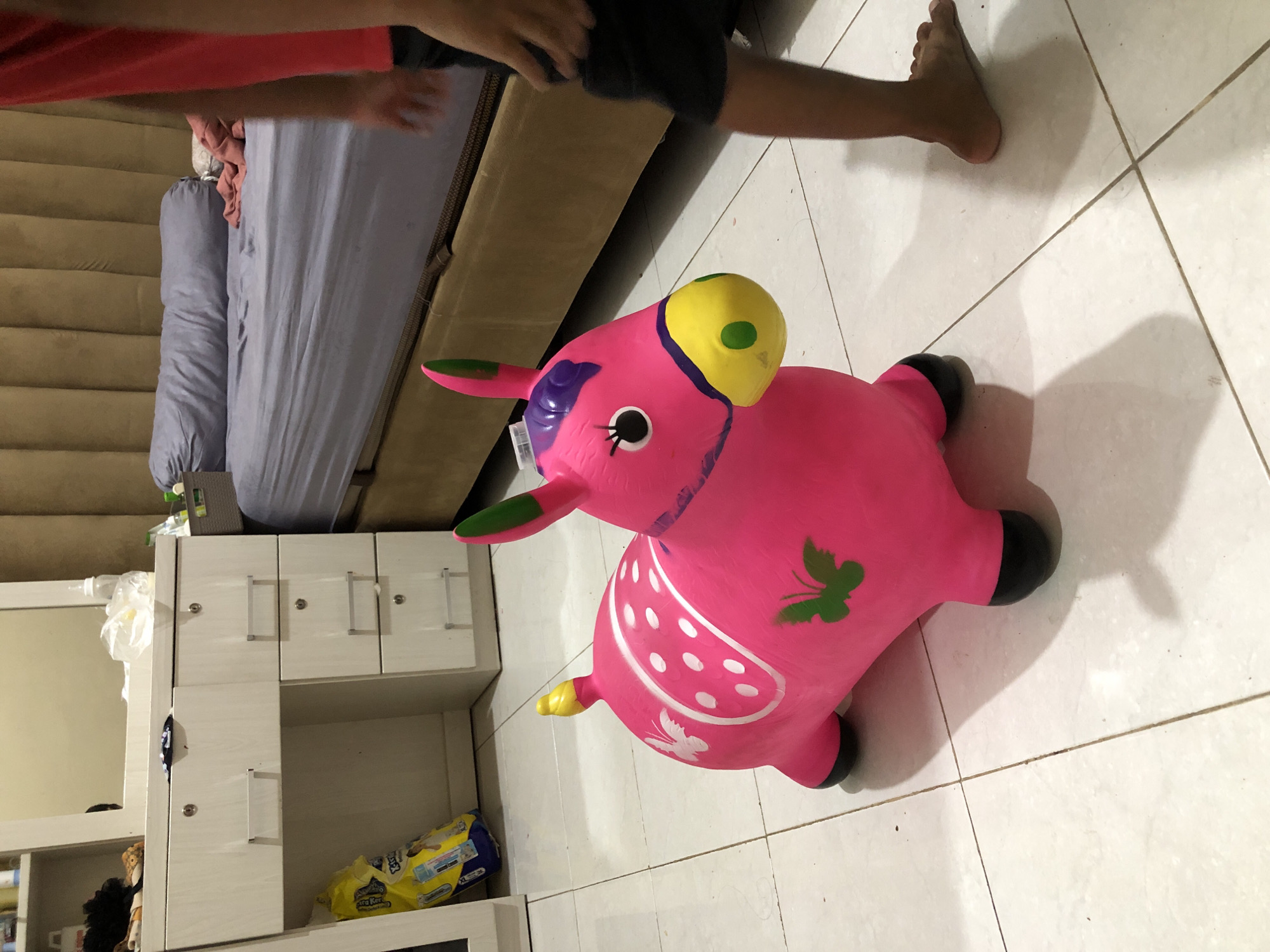 The image depicts an indoor scene, possibly a bedroom or a child's playroom, with a pink inflatable horse toy prominently featured in the foreground. The toy is positioned on a tiled floor. To the left, a white dresser with drawers and a mirror above it is visible. A yellow bag, possibly containing diapers, rests near the dresser. In the background, a bed with a grey headboard and blue bedding is partially visible. A person's legs, wearing dark shorts, are seen on the right side of the image, suggesting their presence in the room. The lighting appears to be artificial, creating shadows on the floor. The overall setting suggests a domestic environment. There is no information or visual evidence to ascertain the location as Pashiebeutong, Indonesia.