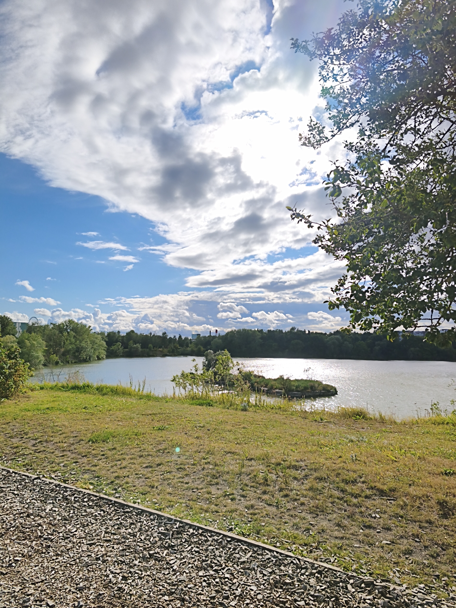 The image depicts a natural scene with a lake in the city of Tallinn, Estonia. In the foreground, a gravel path runs diagonally from the bottom left corner, bordered by a grassy embankment. The embankment is covered with dry grass and sparse green patches, with some scattered debris. Beyond the embankment, a body of water, likely a lake, dominates the midground, reflecting the sunlight. A small, rocky island with vegetation is situated in the lake. The background features a dense line of trees that separates the lake from a distant urban skyline. Silhouetted against the sky are buildings and what appears to be a Ferris wheel, suggesting a recreational area within the city. The sky is filled with a mix of white, fluffy clouds and darker, stormier clouds, with bright sunlight breaking through in the upper right quadrant, casting a lens flare. A tree branch with green leaves extends into the upper right corner of the frame. The overall impression is a blend of natural landscape and urban elements under a dynamic sky.