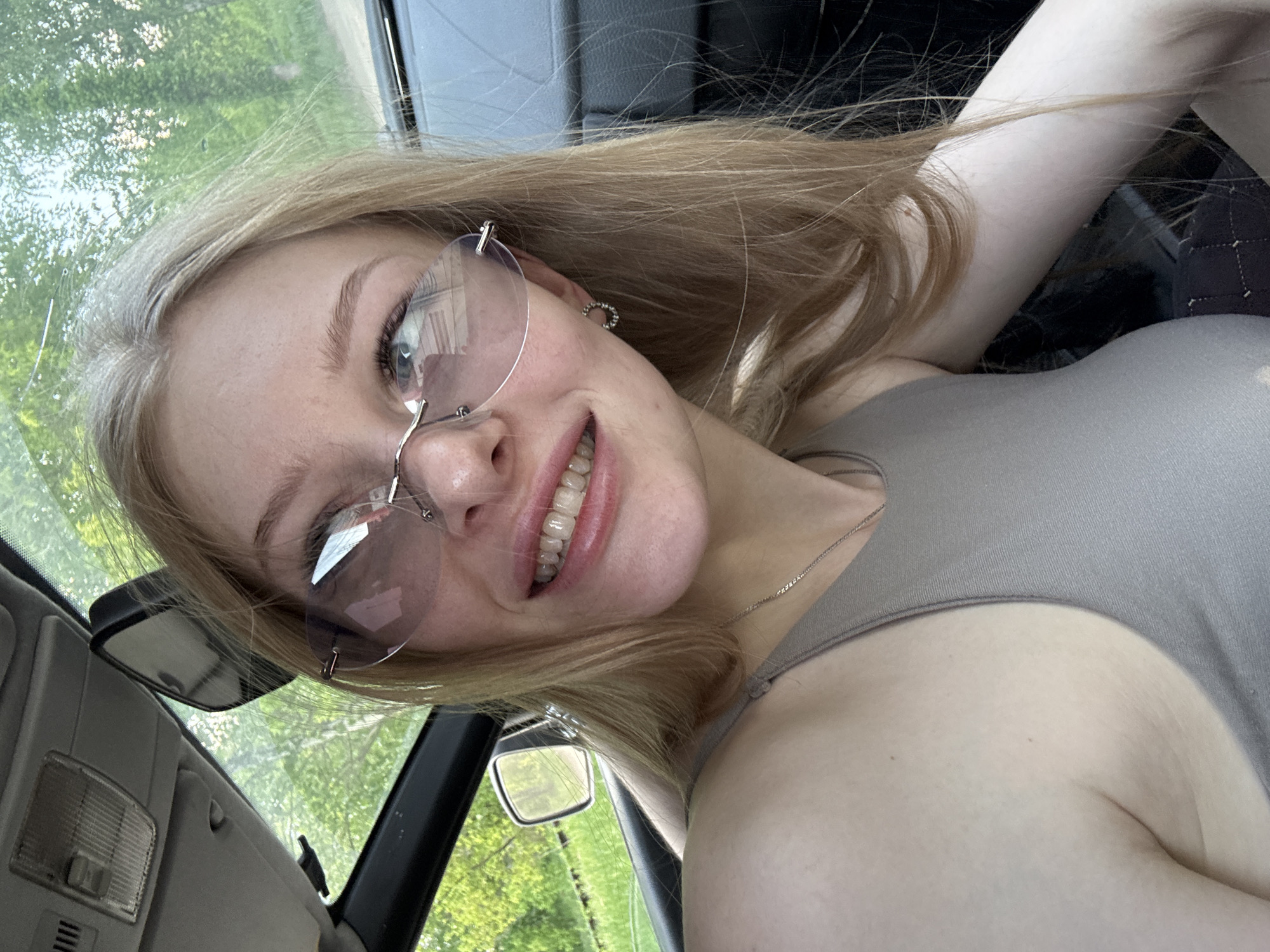 A young woman with light blonde hair and fair skin is positioned inside a car. She is wearing rimless glasses. The car's interior is visible, including parts of the dashboard, a rearview mirror, and the side window. Outside the window, greenery suggests a park or tree-lined street. The scene is possibly located in Perm, Russia, as requested. The woman is smiling and appears to be taking a selfie. She is wearing a grey tank top.