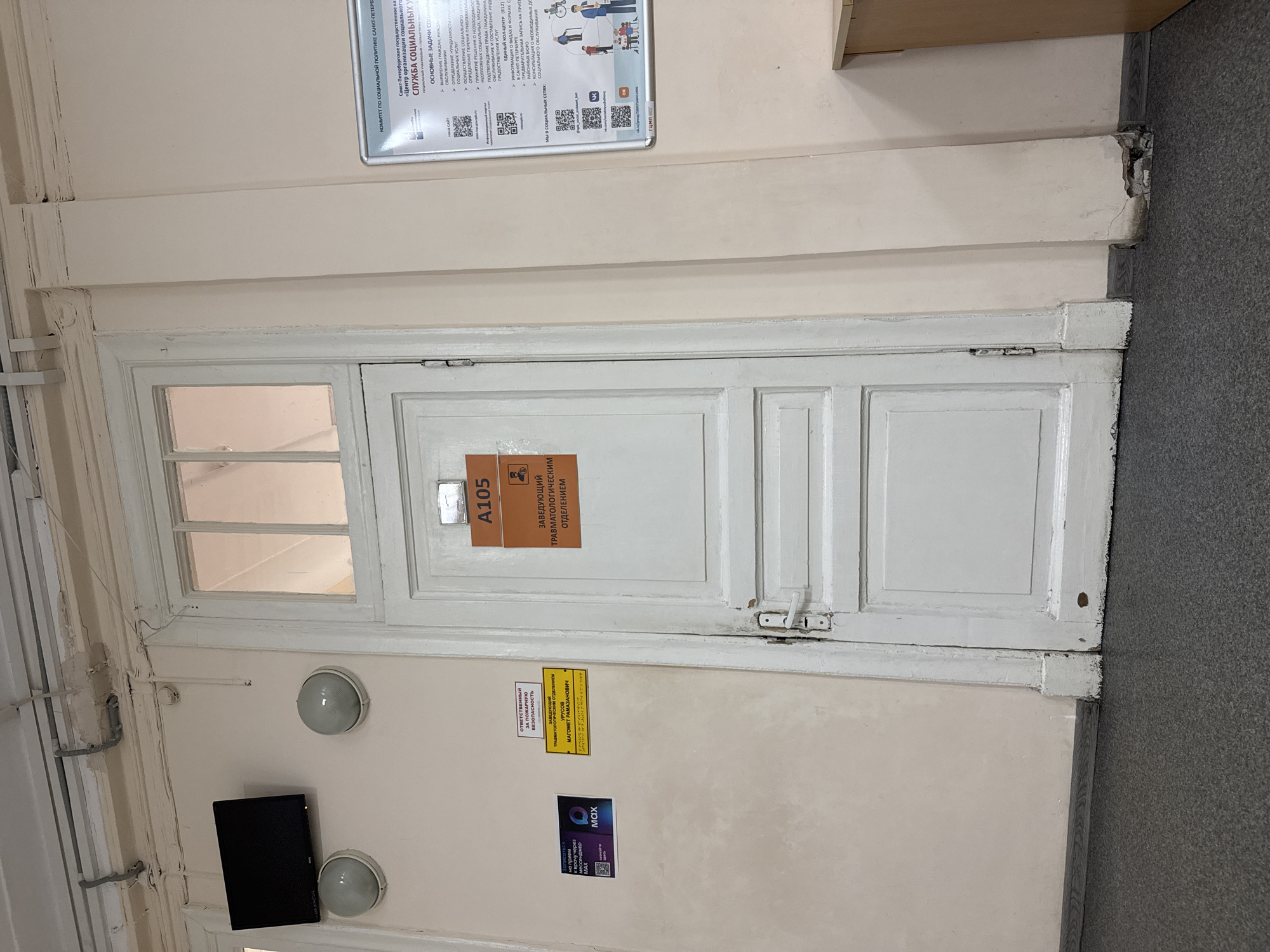 The image shows a hallway with a white wooden door, labeled "A105." The door has a sign that reads "ЗАВЕДУЮЩИЙ ТРАВМАТОЛОГИЧЕСКИМ ОТДЕЛЕНИЕМ" (Head of the Traumatology Department). There is also a smaller sign that says "ОТВЕТСТВЕННЫЙ ЗА ПОЖАРНУЮ БЕЗОПАСНОСТЬ" (Responsible for Fire Safety). On the wall to the left of the door, there is a black television screen mounted above two spherical lights. Further to the left, there is an advertisement poster with a QR code. A bulletin board with text and QR codes is mounted on the wall to the right of the door, featuring information about the "Committee for Social Policy of Saint Petersburg" and the "Social Services Center." The hallway floor is carpeted in dark gray, and the walls are painted a light beige. The overall condition of the hallway appears to be dated, with some wear and tear visible on the doorframe and wall. The lighting in the hallway is somewhat dim.