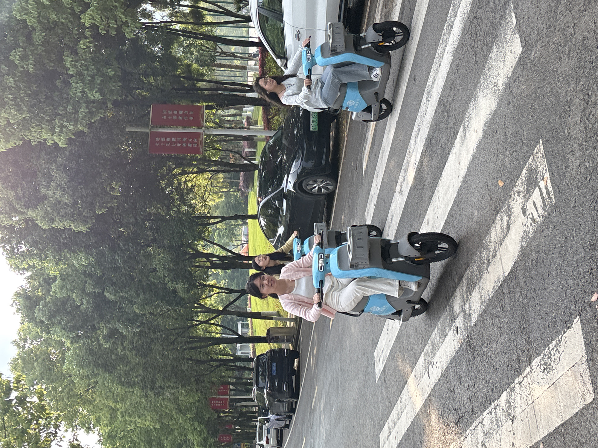 The image shows three young women riding blue-and-grey electric scooters across a zebra crossing. They are traveling in the same direction. Cars are parked along the side of the road. A red sign with Chinese characters is visible. Trees line the road, with green foliage. The license plate "MA DL328" is visible on one of the vehicles. Based on the information and image analysis, the location is in Jiangjin, China.
