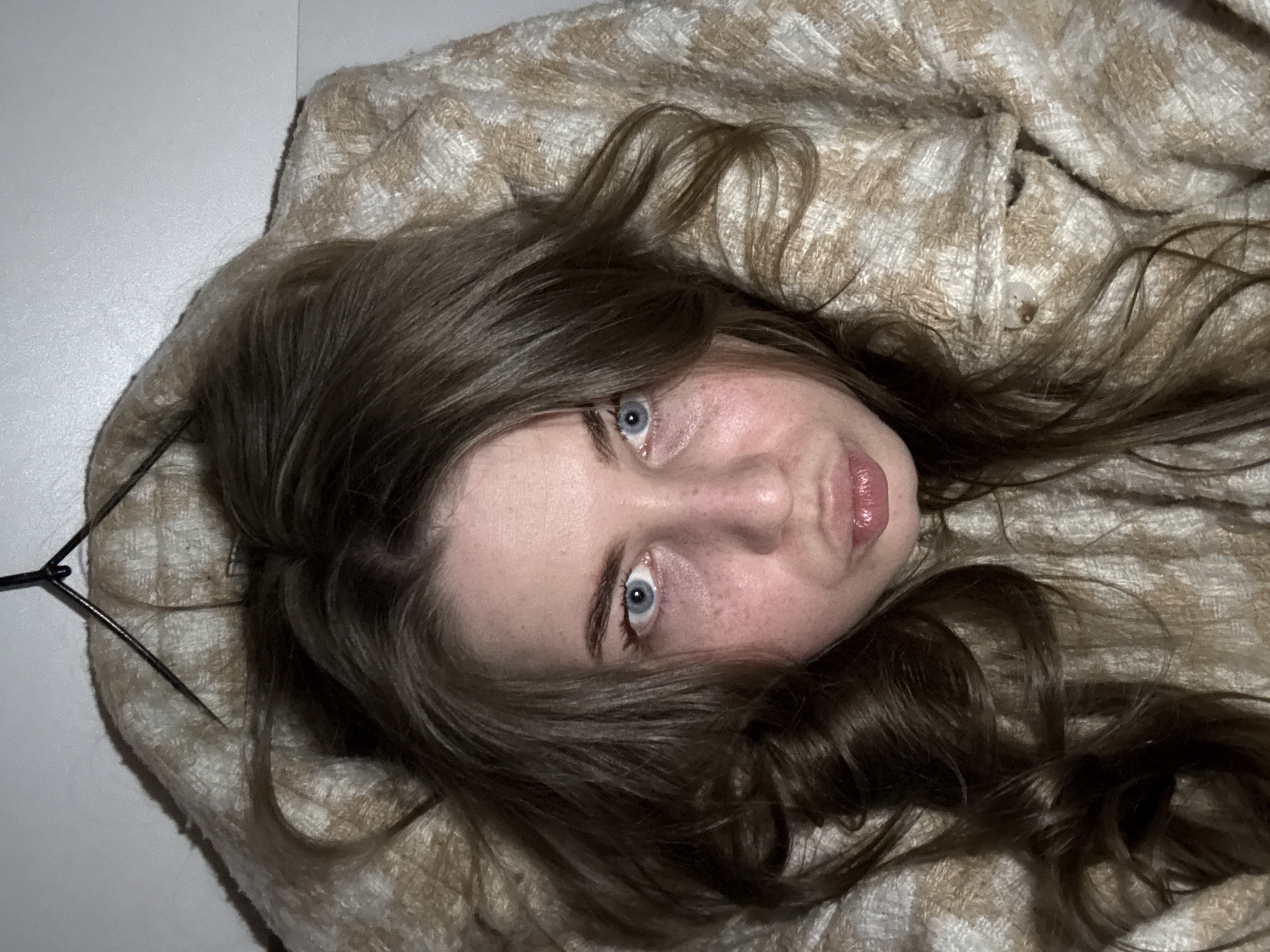 A young woman is depicted in a close-up, overhead shot, lying down with her head rotated approximately 90 degrees clockwise relative to the frame, looking directly upwards at the camera. Her long, wavy, dark brown hair cascades around her head and shoulders, spreading across a textured fabric.
Her bright blue eyes are wide open, engaging directly with the viewer. Her full lips are slightly pursed with a glossy finish, contributing to an expression that appears curious, thoughtful, or playfully pouty. Her fair skin shows a scattering of faint freckles across her cheeks.
She is resting on a soft, woven fabric, possibly a blanket or a jacket, which features a repeating pattern of light brown, beige, and off-white squares or checks. In the upper left portion of the image, a plain, light grey or off-white wall serves as the background. A thin, dark, wire-like object, possibly the arm of eyeglasses or a clothes hanger, is partially visible near her head on the left side of the frame.
The lighting is soft and diffuse, highlighting her features without harsh shadows. The image captures an intimate, candid moment. No text is visible in the frame. The location context for this image is Chaikovsky, Russia.