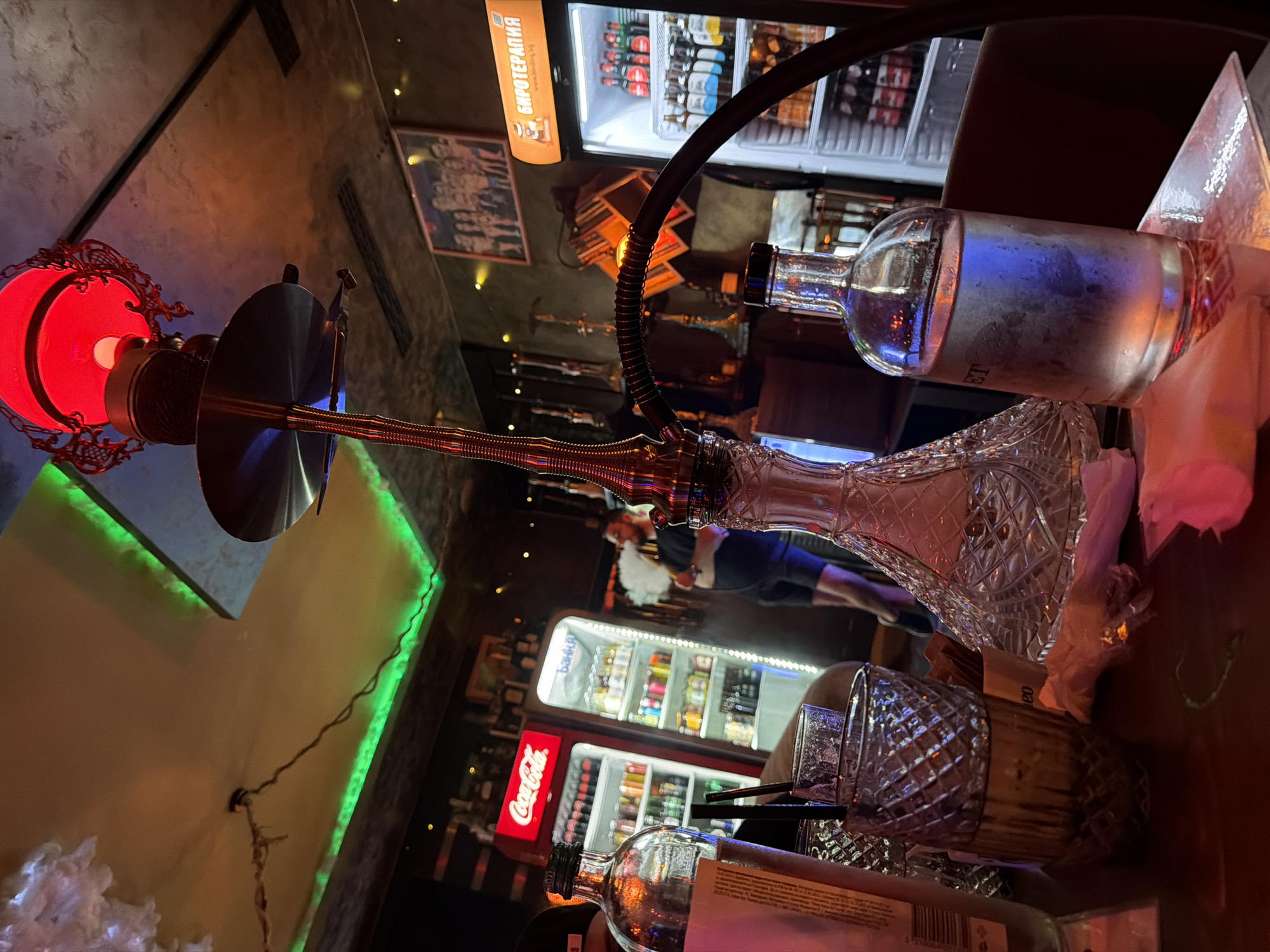 This image, tilted to the right, depicts an indoor scene in what appears to be a dimly lit bar, lounge, or hookah establishment. The primary subjects are a hookah, various glass bottles, and tumblers on a wooden table, set against a backdrop of refrigerators and textured walls with accent lighting.
A prominent hookah stands on the table, featuring a red illuminated bowl, a metallic stem, a black flexible hose, and an intricately patterned clear glass base. To the right of the hookah, a frosted glass bottle, likely containing alcohol, rests on its side. Another clear glass bottle with a label and barcode is visible at the bottom of the frame, alongside several patterned glass tumblers with black straws. Paper napkins or tissues are scattered on the table surface.
In the background, two refrigerators are visible. The top one, brightly lit, is stocked with various bottled beverages and features a sign with the Cyrillic text "БИРОТЕРАПИЯ" (BIROTERAPIYA), and part of a website address "www.bir... .bg". Below it, a red Coca-Cola branded refrigerator is also filled with drinks. A person's arm or shoulder is partially seen near the Coca-Cola fridge, and another person's leg or torso is subtly visible further back.
The setting is characterized by low, ambient lighting, with a green LED strip providing accent light along a wall. A framed poster or picture hangs on the wall near the top refrigerator. The time of day is indicated as evening or night due to the reliance on artificial lighting. The overall atmosphere suggests a casual social gathering.
