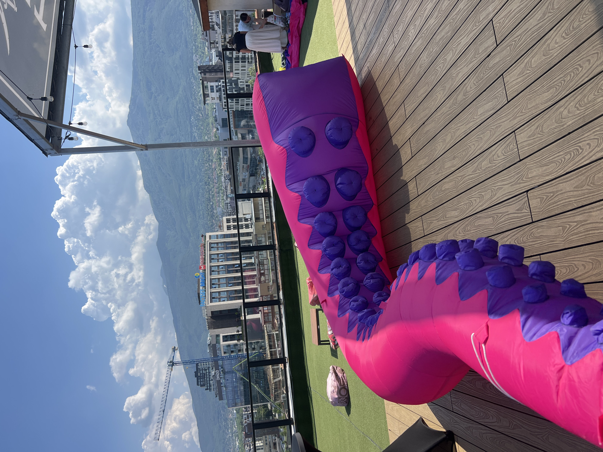 The image shows a rooftop in Sofia, Bulgaria, on a sunny day with scattered clouds. In the foreground, a large, pink and purple inflatable tentacle is laid out on a wooden deck. The tentacle has purple circular suction cups running along its length. To the left, there is artificial turf with a small wooden table and a pink beanbag. In the background, several modern buildings and a construction crane are visible, with mountains in the distance. Two people, a woman in a white skirt and a man in a blue shirt, are tending to the inflatable structure. The scene suggests an outdoor event or installation.