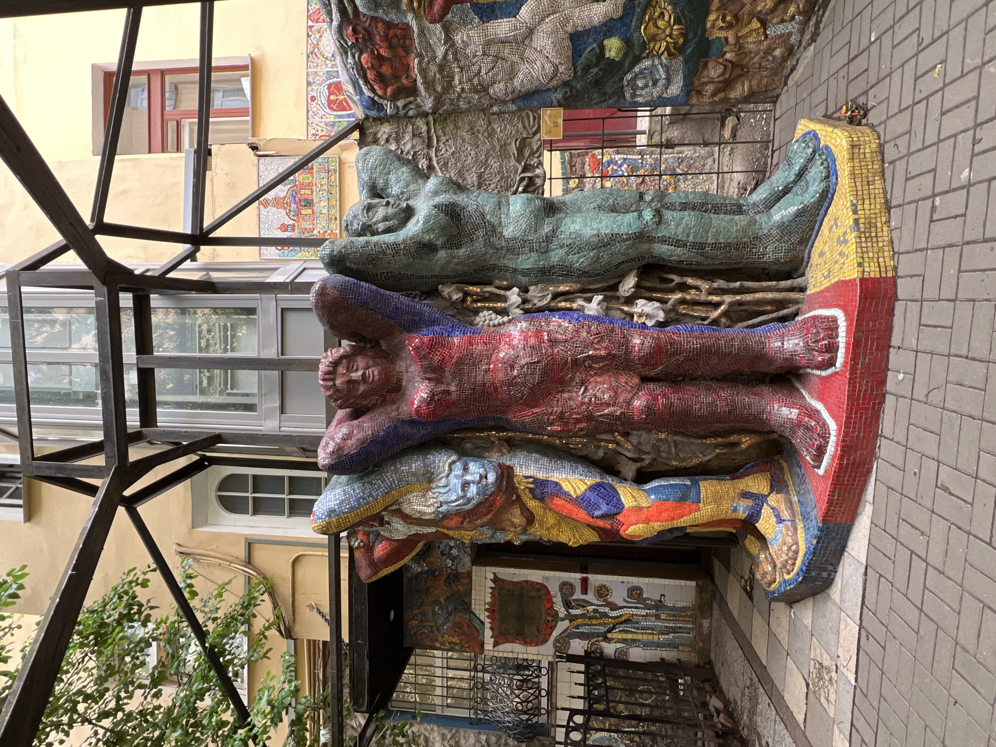 This media file captures an outdoor scene featuring a mosaic art installation of figures situated against a building in Saint Petersburg, Russia. The artwork consists of three layered, stylized human figures, each rendered in vibrant mosaic tiles. The topmost figure appears to be lying on its back, with its torso and limbs visible. Below this, a red-tiled figure is depicted in a similar reclined pose. The lowest visible figure is also tiled, with its head and upper body showing. The mosaic style is detailed, with many small tiles forming the contours and textures of the figures.
The setting includes the facade of a building with yellow paint and architectural elements like windows. One window has a red frame, and another is a round-topped window with multiple panes. A dark metal structure, possibly a balcony or staircase support, is prominent in the foreground and partially obscures the view. Green foliage from a tree or bush is visible on the left side of the frame, reaching towards the metal structure. The ground is paved with light-colored rectangular tiles. The lighting suggests it is daytime, and the weather appears to be overcast or at least not sunny, given the even illumination and lack of strong shadows. The overall impression is one of an art installation integrated into an urban environment, possibly a courtyard or a side street. There is no visible text in the image.