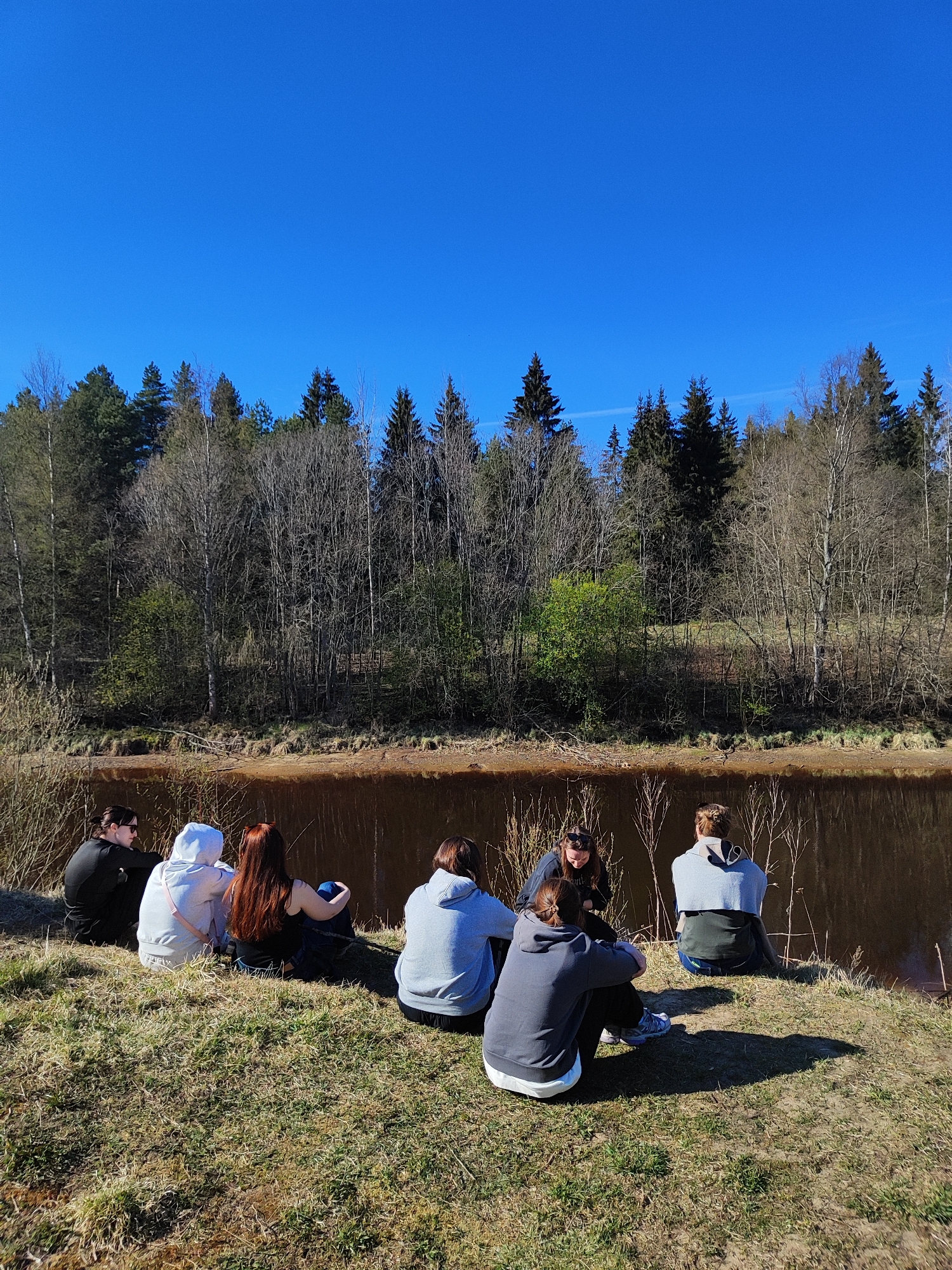 A group of seven people are sitting on a grassy riverbank, their backs facing the viewer. The people are of varying builds and clothing styles. The background is a line of trees and bushes separating the river from the sky. The sky is clear and blue. The location is the city of Unknown, Unknown.
