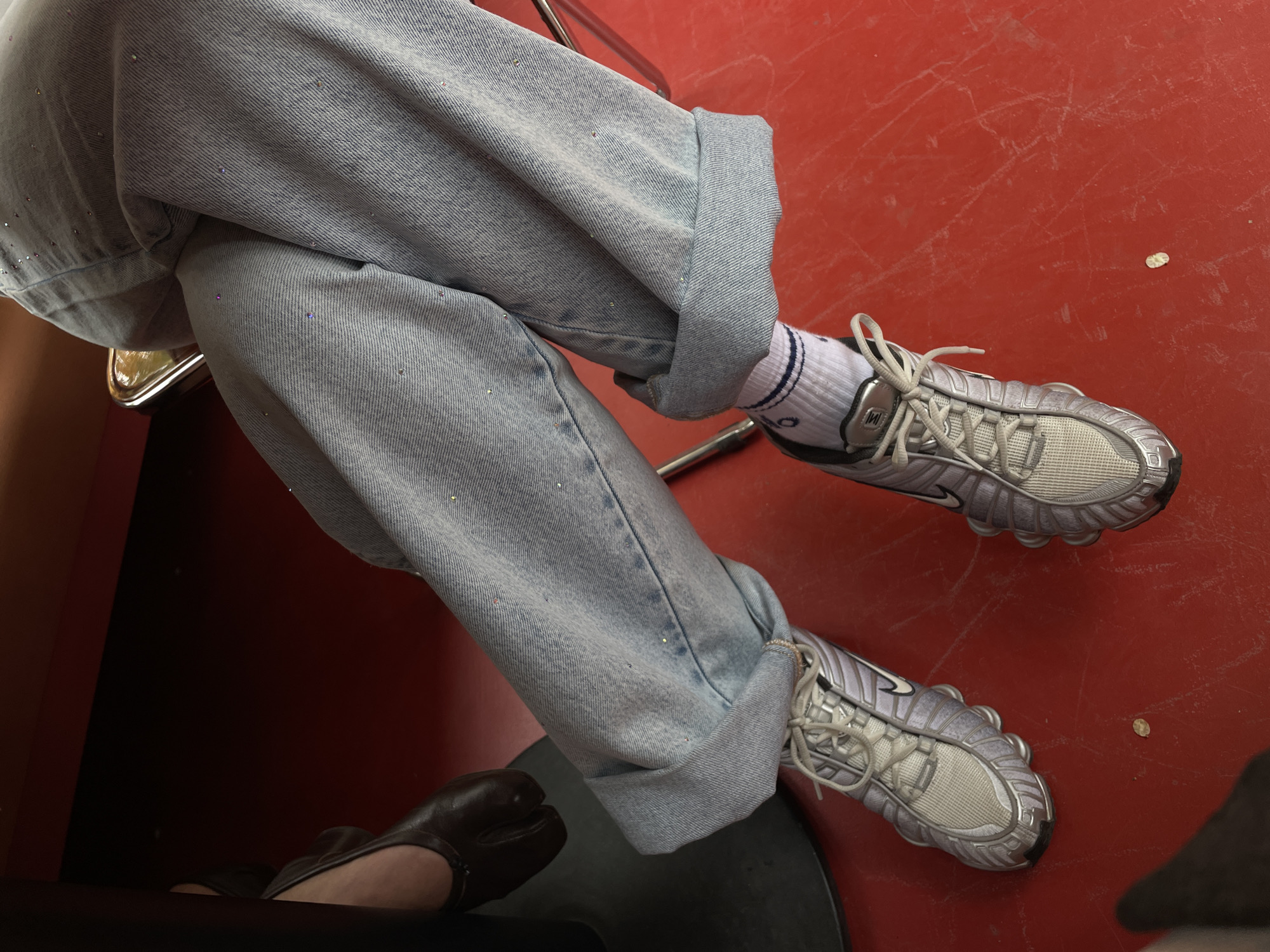 A close-up shot from above shows a person sitting with their legs crossed against a red background. The person wears light blue, embellished denim jeans with the cuffs rolled up and white socks with a navy blue stripe near the top. They are wearing gray athletic shoes. The toes of a brown leather shoe peek out from under the jeans. The location could be Yerevan, Armenia, but there are no specific elements in the picture to confirm.