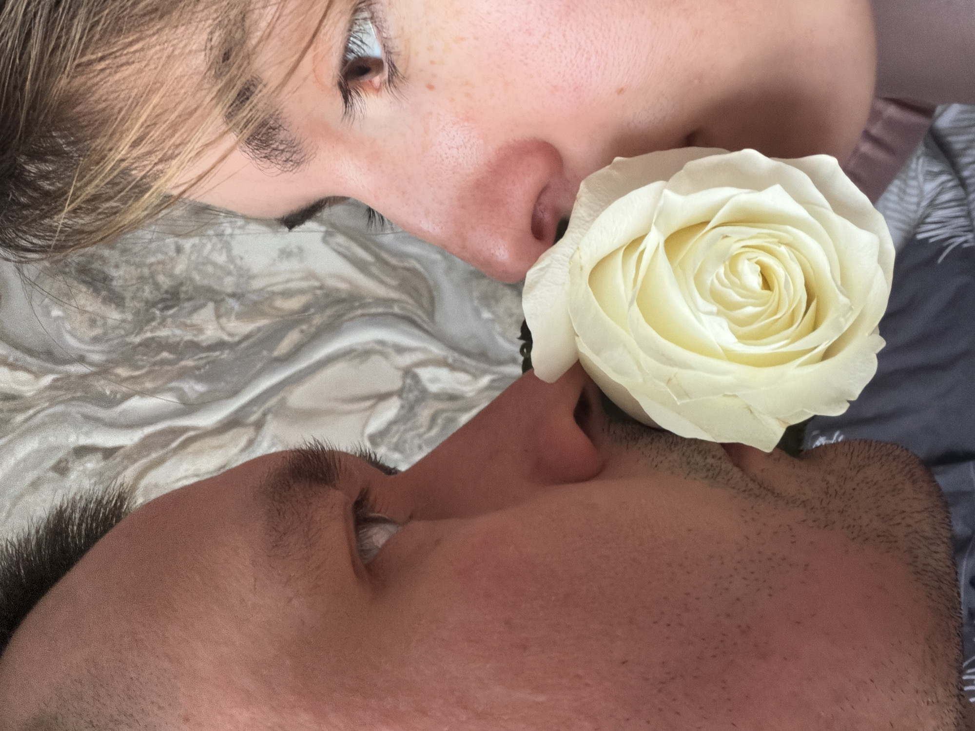 A close-up shot features two individuals, a fair-skinned woman and a darker-skinned man, positioned face-to-face with a white rose placed between their noses. The woman is located at the upper part of the image, and the man is positioned below. Both individuals are lying on a textured surface with silver/gray-colored cloth behind them. The location is not identifiable from the image.
