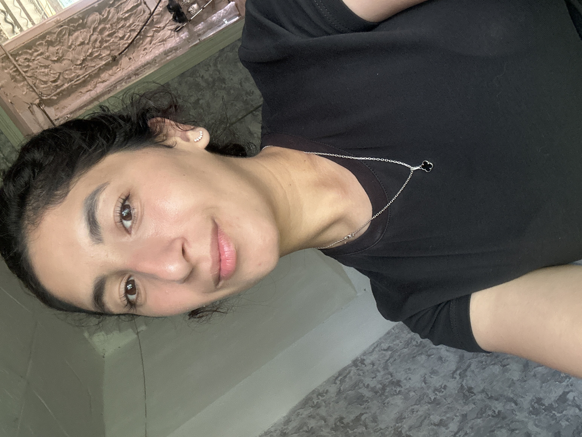 A young woman with dark hair pulled back and brown eyes is looking at the camera. She is wearing a black t-shirt and a silver necklace with a clover pendant. She has small, delicate earrings in her right ear. The background appears to be indoors, with a textured gray wall on the left and a lighter, possibly stuccoed wall on the right. Some architectural details are visible in the upper right corner. There is no indication of the city of Dondukovskaya, Russia in the image.