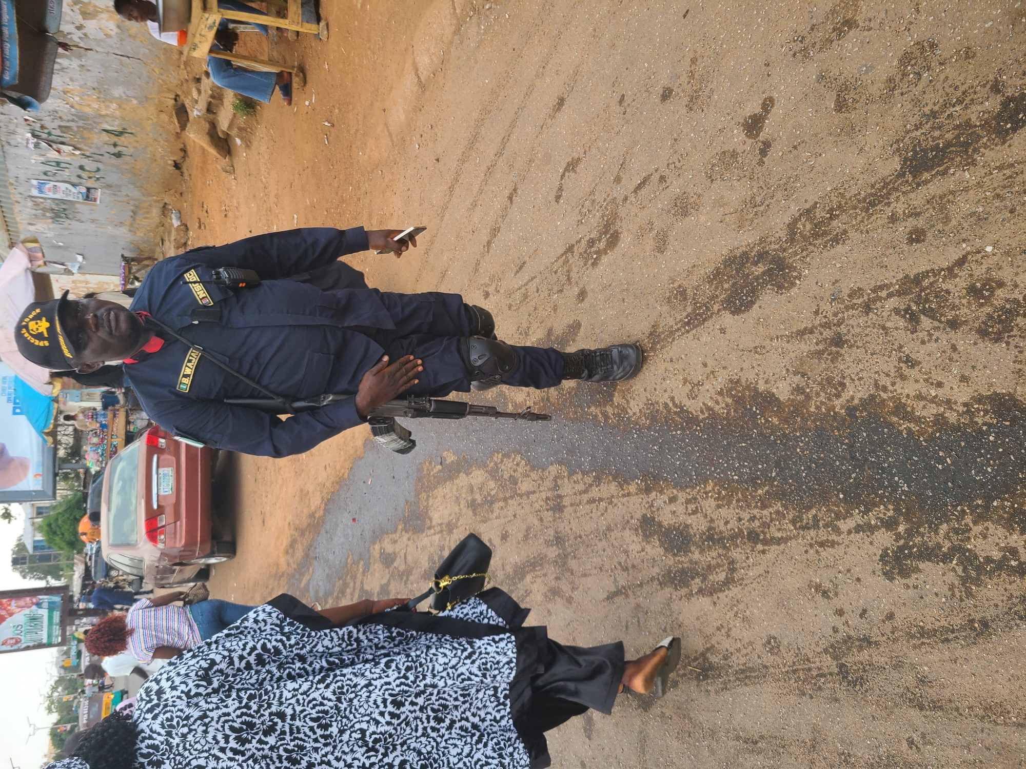 The image depicts a man in dark blue tactical gear, identified by patches as a member of S.W.A.T. (Special Weapons and Tactics), standing on a street in Jos, Nigeria. He is holding a rifle and a cell phone. A woman in a black and white patterned garment is walking by. The street appears to be paved but has patches of dirt and dark liquid stains. A maroon sedan is parked nearby. There is signage in the background indicating commercial activity and other people are visible further down the street.