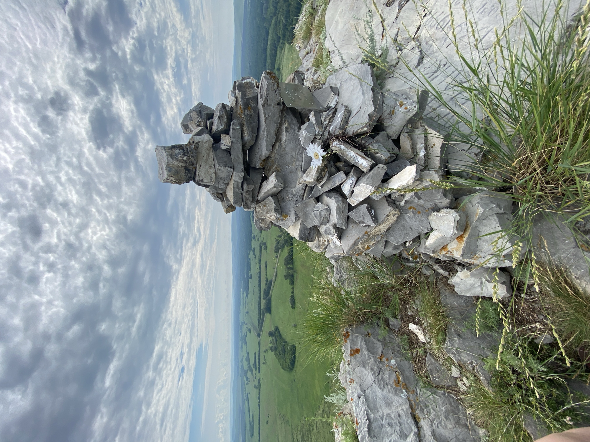 A cairn of gray, irregular stones is stacked on a rocky outcropping. A white daisy is placed on the stones. A metal plaque is affixed to the cairn. Green grass and vegetation grow around the stones. In the background, a vast, rolling green landscape stretches to the horizon under a cloudy sky. The scene appears to be in a natural, elevated area, possibly a viewpoint.
