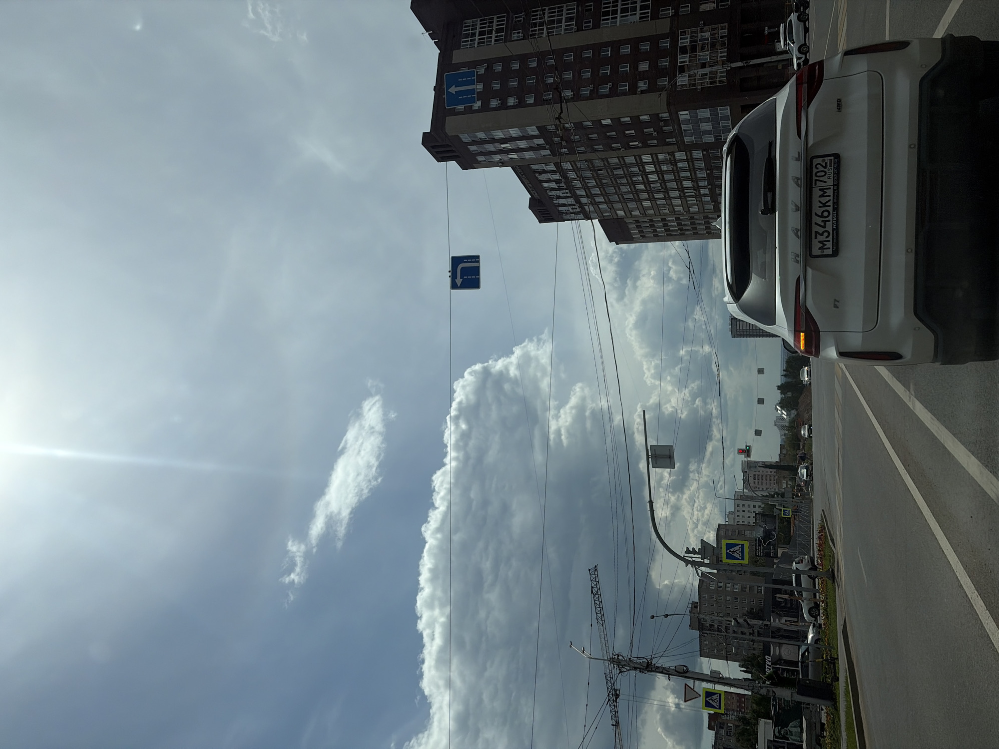 The image captures a street scene in Ufa, Russia, under a partly cloudy sky. A white Haval SUV is prominently featured, its rear end facing the viewer. The car's license plate displays the text "M346KM702". In the background, a tall, multi-story brick building with numerous windows dominates the skyline. Several road signs are visible, including a blue sign with a white left arrow and another blue sign with a white right arrow. The sun is shining brightly, casting a lens flare on the left side of the image. The street appears to be an urban area with other vehicles and pedestrian crossings visible further down the road. The overall impression is a snapshot of everyday life on a city street.