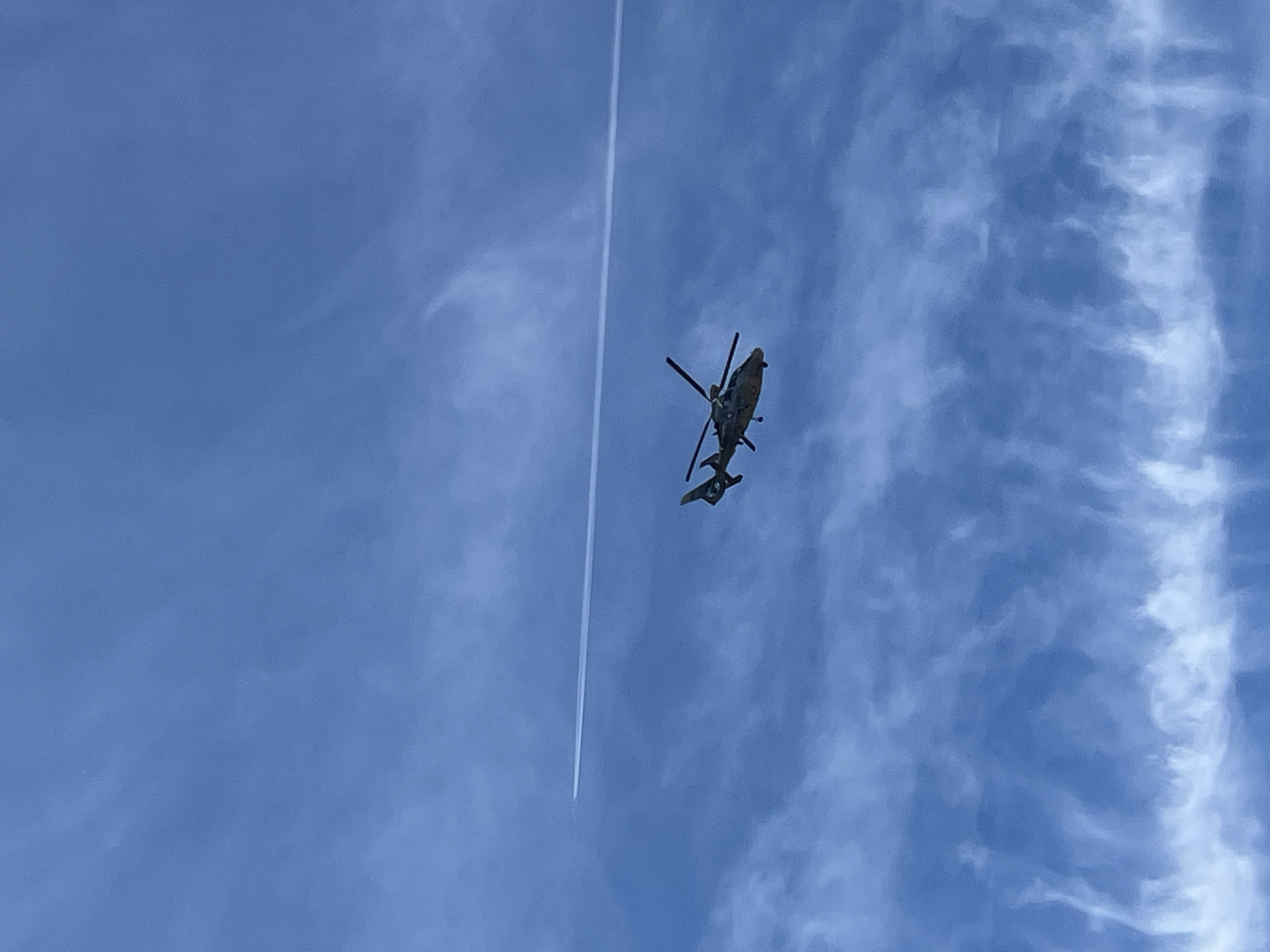 A helicopter is airborne in a blue sky with cirrus clouds. A white contrail is visible near the helicopter. The location is likely Pchelnik, Bulgaria, given the prompt.