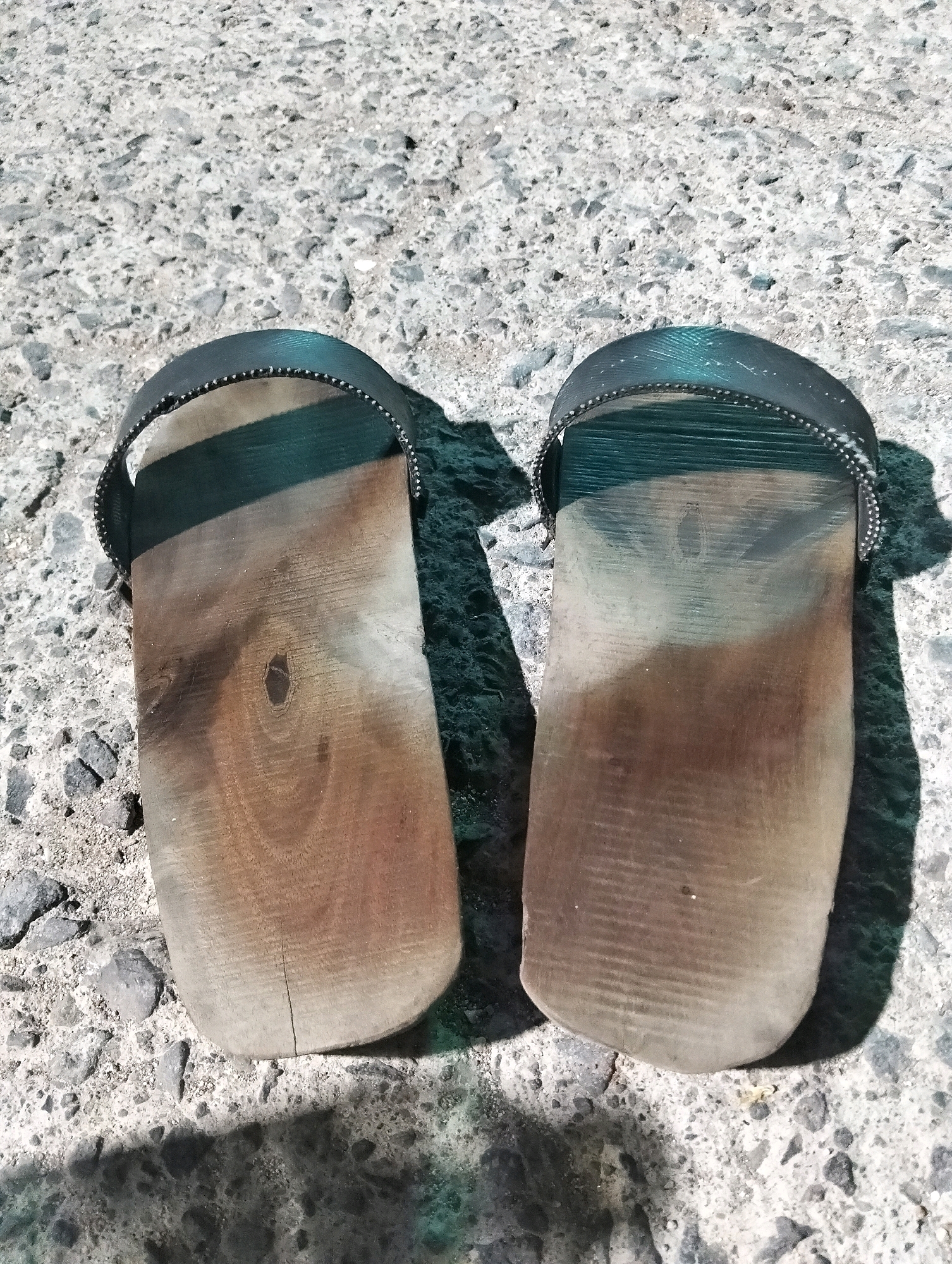 This is a top-down, close-up, eye-level photograph of a pair of wooden Japanese Geta sandals resting on a gravel surface. The sandals are made of light-colored wood with a dark, textured strap that goes over the foot, secured by small studs. The wood is worn, showing grain patterns and some discoloration. One sandal has a small crack near the toe. The gravel surface is a mix of grey and white stones with visible dirt. Shadows of the sandals are cast on the gravel, indicating direct sunlight from the left. The scene appears to be outdoors, possibly a pathway or a yard. There are no people or animals visible in the image. The location is identified as Paciran, Indonesia.