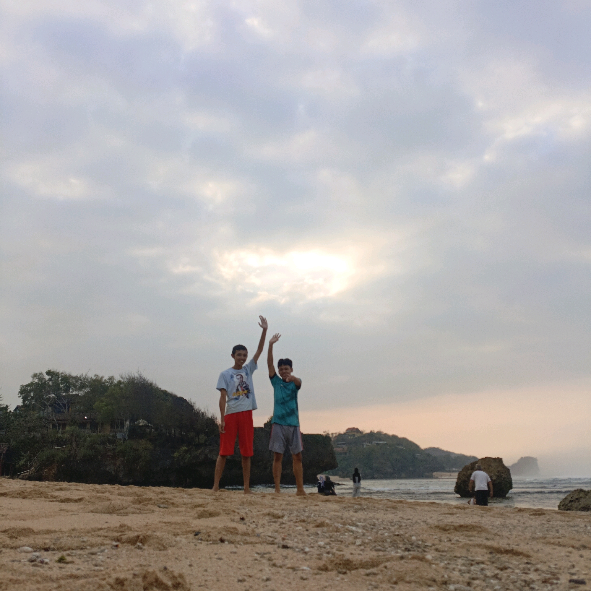 Two young males stand on a sandy beach in Tepus, Indonesia, with their arms raised in a cheerful gesture. The male on the left wears a white t-shirt and red shorts, while the male on the right is dressed in a teal t-shirt and gray shorts. They are positioned in the foreground of the image. In the background, a body of water meets the shoreline, with a rugged coastline and vegetation forming a backdrop. The sky above is overcast, with the sun partially visible through a break in the clouds, casting a soft light on the scene. A few other individuals and a large rock are visible further down the beach. The overall atmosphere suggests a casual outing at the beach.