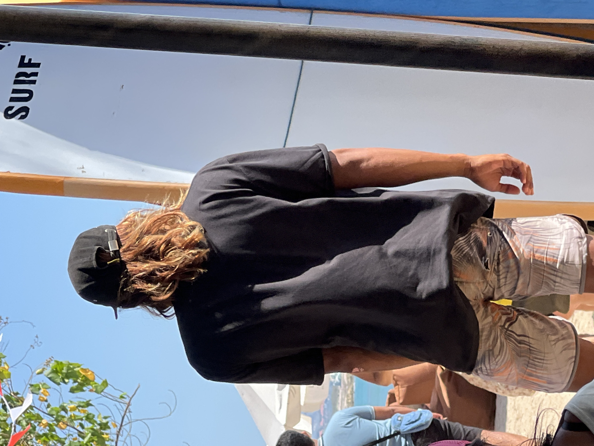 In Kuta, Indonesia, on a bright, sunny day, a person stands with their back to the camera. They are wearing a black t-shirt and patterned shorts, with a black baseball cap on their head. Their long, reddish-brown hair is visible beneath the cap. To their right, a white surfboard with the word "SURF" in black lettering stands upright, leaning against a pole. The background shows a clear blue sky, with hints of green foliage from a tree on the left and a portion of a building or structure to the far left, partially obscured by an umbrella. Several other people are visible in the background, including one person in a light blue shirt and another figure sitting down. The scene appears to be on a beach or in a beachside area, suggested by the sand and casual attire.