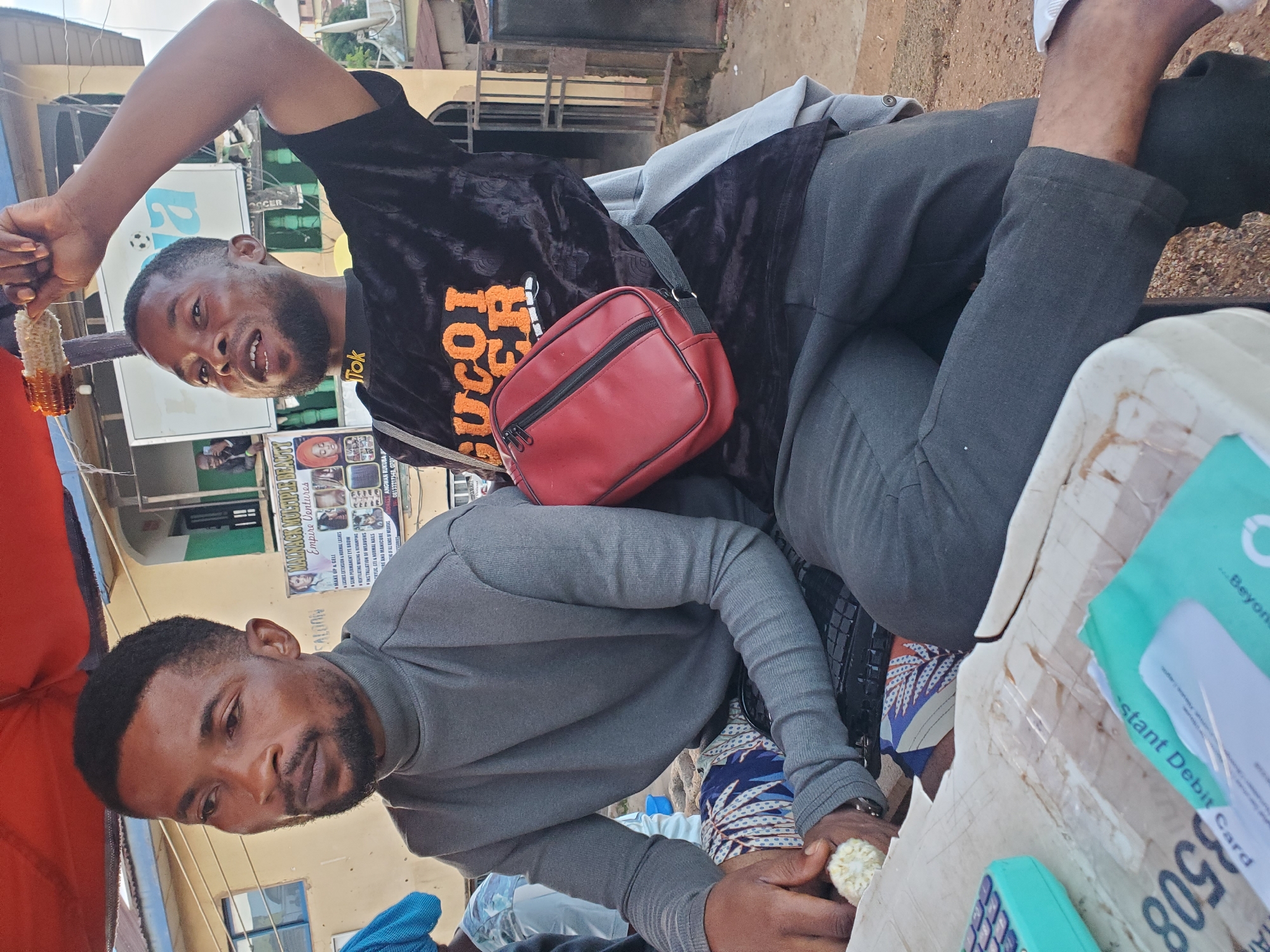 In Gwong, Nigeria, two young Black men are seated outdoors under a red awning. The man on the left wears a grey turtleneck sweater and patterned trousers. He holds a piece of corn in his left hand and a calculator in his right. The man on the right wears a black shirt with orange lettering and dark pants. He raises a piece of corn in his right hand and is looking up and to his left. A red bag is slung across his body. In the background, a building with signs and a metal gate is visible. The ground is covered in dirt and gravel.