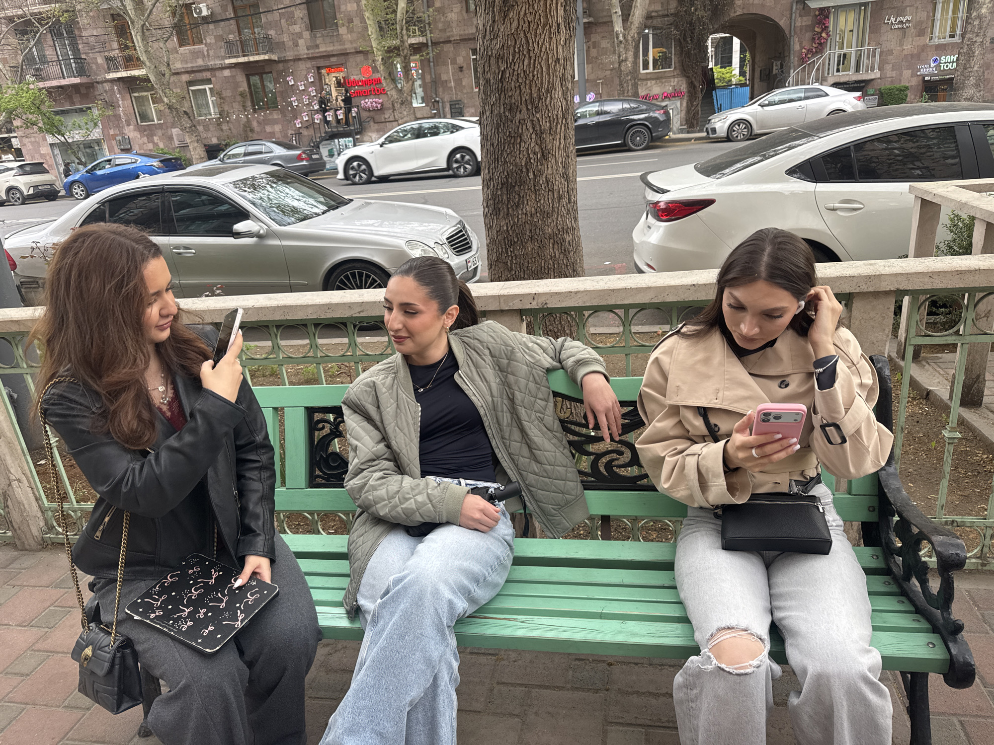 Three young women are seated on a green park bench in an urban setting, likely Yerevan, Armenia. The scene appears to be daytime under an overcast or partly cloudy sky.
The woman on the left, with long brown hair, wears a black leather jacket over a dark top and dark trousers. She holds a smartphone in her left hand and looks towards the middle woman. A black cross-body bag with a chain strap is visible, and she has a black notebook or tablet with a white patterned design on her lap.
The middle woman, with her dark hair pulled back, wears a black top under a quilted light green bomber jacket and light wash baggy jeans. She is looking towards the woman on her left with a slight smile. A black fanny pack is visible around her waist.
The woman on the right, with dark hair, wears a beige trench coat over a dark top and light wash ripped jeans. She is focused on a pink-cased smartphone in her hands and has white earbuds in her ears, one of which she is adjusting. She also has a black fanny pack around her waist.
Behind the women, a city street with cars is visible. Parked and moving vehicles include a silver Mercedes-Benz E-Class, a white Mazda 6, a blue sedan, and other cars. Multi-story stone buildings with balconies and windows line the street. Large tree trunks separate the bench area from the road.
Visible text on storefront signs includes "Xiaomi" and Armenian words such as "Սմարթֆոն" (Smartphone) and "Լինիա Տուր" (Linia Tour) with its website "www.liniatour.am". The overall mood is relaxed and casual, suggesting the women are socializing or passing time.