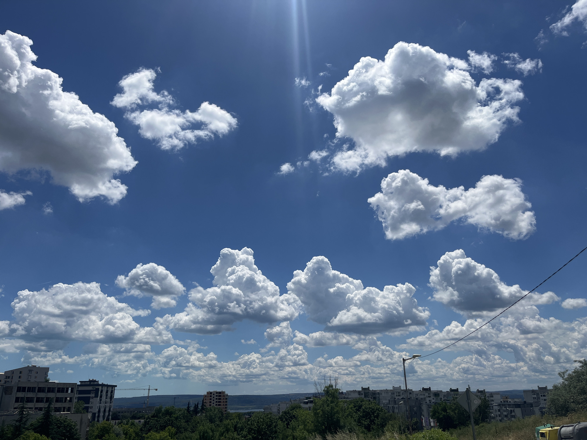 The image shows a view of the city of Varna, Bulgaria, from an elevated position, looking towards the east. The sky is a vibrant blue, filled with fluffy white cumulus clouds. The sun is high in the sky, casting bright light and creating visible sunbeams. In the foreground, there is a lush green area with tall grass and some sparse bushes.
Dominating the middle ground and stretching into the background are residential buildings, typical of Soviet-era architecture, with multiple stories and a uniform design. Some of these buildings appear to be in various states of repair, with some showing signs of wear. A construction crane is visible in the distance, suggesting ongoing development or renovation in the city. The buildings are interspersed with trees, forming a dense green canopy.
The horizon line is visible, separating the city from the sky. Further in the distance, the landscape appears to be a mix of urban structures and natural greenery, with a hint of water, possibly the Black Sea, in the far distance. A single street lamp and a traffic sign are also visible in the foreground, indicating the presence of infrastructure. The overall scene depicts a bright, sunny day over a residential area of Varna.