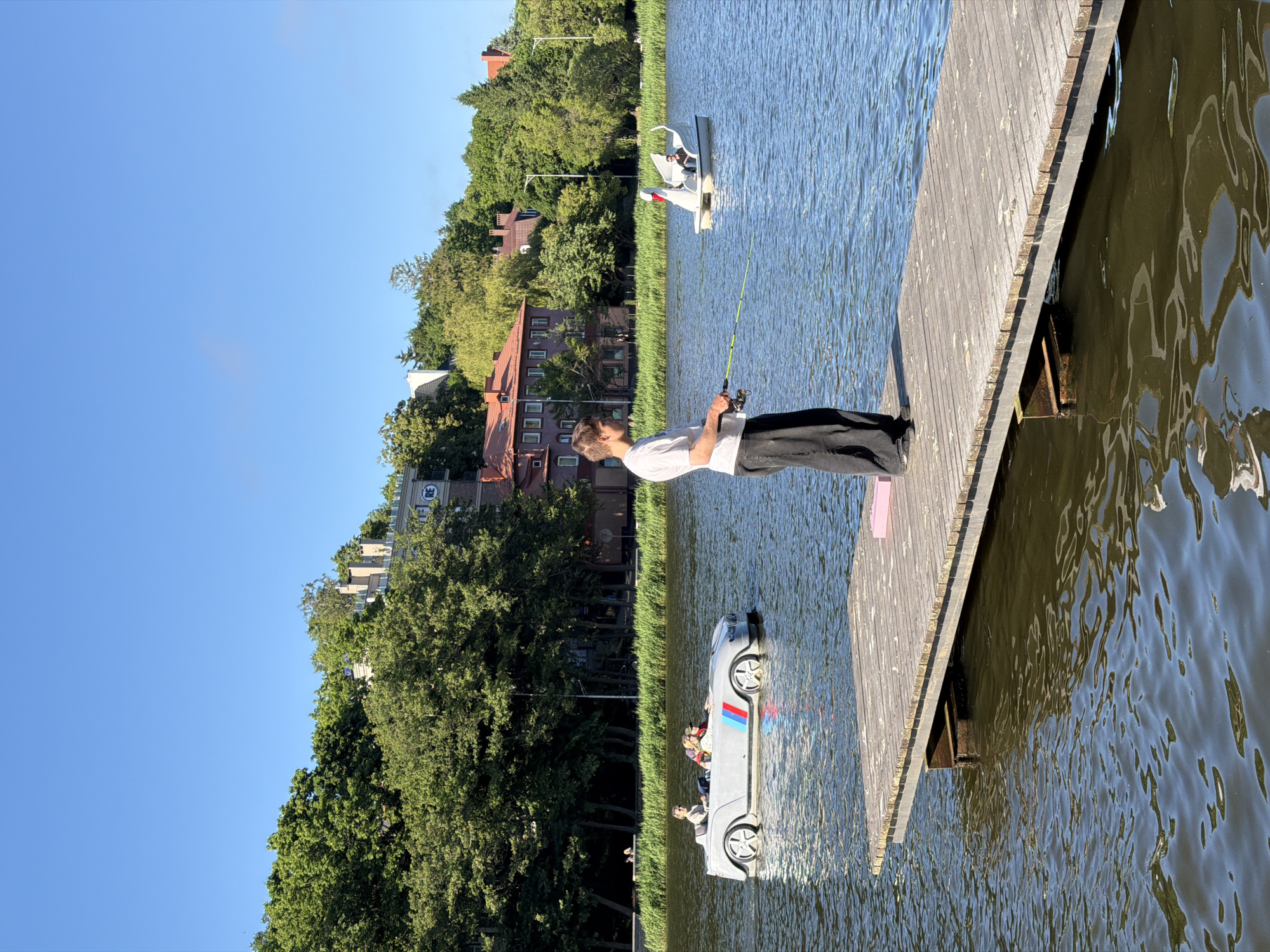 A young man stands on a wooden pier, fishing with a rod in a body of water. He is wearing a white t-shirt and black pants. The pier extends into the water, which is calm and reflects the clear blue sky. In the background, a swan-shaped pedal boat with two people is visible, as well as a grey car-shaped pedal boat with three people. Behind the water, a lush green hillside is covered with trees and buildings, including houses with red tile roofs. The scene appears to be set in a park or recreational area. The location is the lake near the city of Svetlogorsk, Russia.