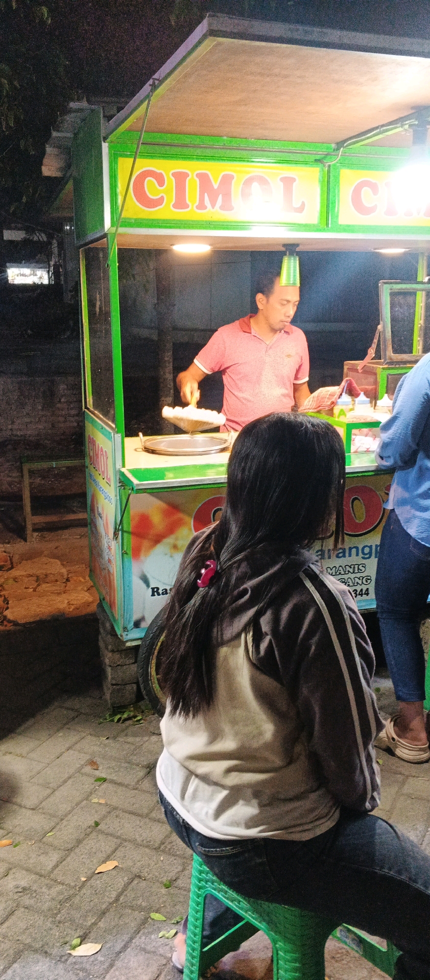 A street vendor in Karangploso, Indonesia, is seen preparing food at a brightly lit stall. The vendor, a man wearing a pink polo shirt, is focused on his task, holding a tool over a circular cooking surface. Above him, a green plastic cup is balanced precariously on his head. The stall has a green frame and a yellow banner with orange lettering that appears to say "CIMOL CIMOL." A young woman with long dark hair, wearing a grey hoodie and dark jeans, sits on a green plastic stool facing away from the camera, observing the vendor. Another person, wearing a blue shirt and blue jeans, stands to the right of the stall. The ground is paved with interlocking bricks, and fallen leaves are scattered around. The background is dark, suggesting it is nighttime, with some indication of trees and a brick wall.