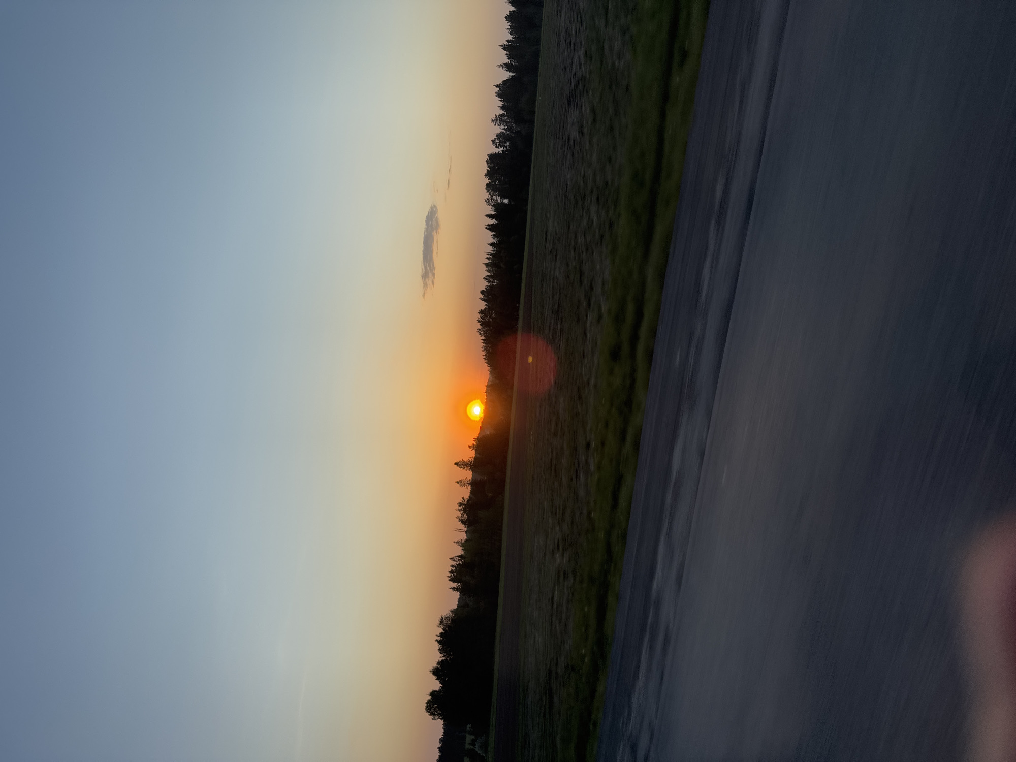 The image depicts a sunset over a field, likely taken from a moving vehicle. A road occupies the right side of the frame, its surface appearing wet. Beyond the field, a line of trees separates the sky from the ground. The sun is positioned near the horizon, casting a warm glow and creating a lens flare. A faint cloud is visible in the upper portion of the sky. The color gradient in the sky transitions from blue to orange.
There is no information to associate the image with Kamennoye, Russia.
