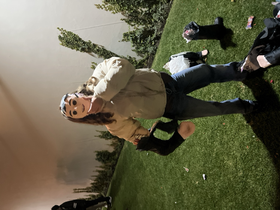 A person wearing a mask, jeans, and a jacket stands on green grass. Near the person are remnants of a smashed piñata, including a black cutout and candy wrappers. A white wall with climbing vines is visible in the background, and the sky is a hazy orange color. Another individual is partially visible near the bottom of the frame.