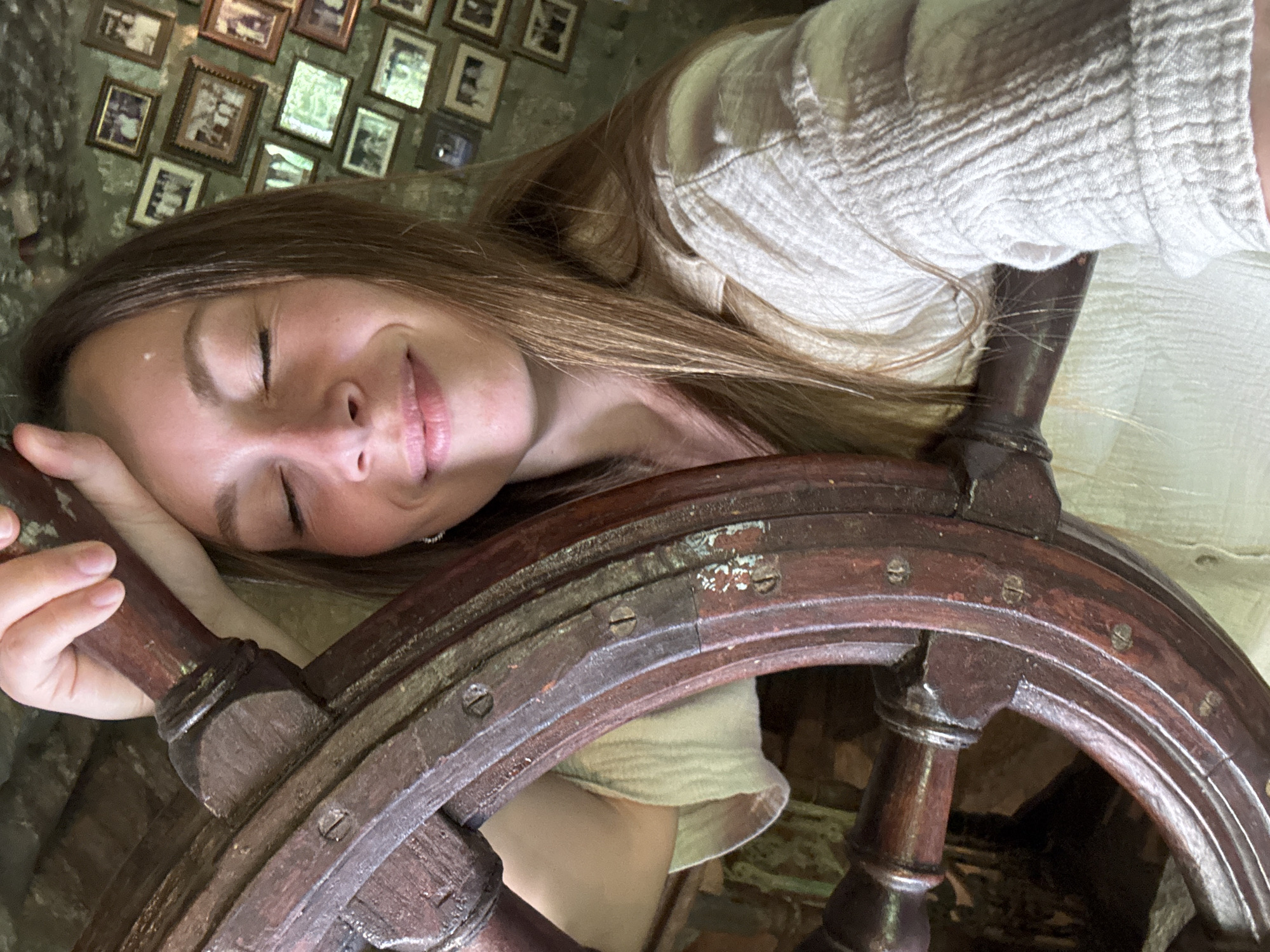 A young woman with her eyes closed and a slight smile is leaning her head against a wooden ship's wheel. She has long, light brown hair and is wearing a light-colored, textured shirt. The ship's wheel appears old and weathered, made of dark wood with metal accents. Behind her, a wall is covered with numerous framed photographs, many of which appear to be vintage family portraits. The setting suggests an indoor location with a rustic or historical theme, possibly a museum or a decorated private residence in the town of Morinj, Montenegro.