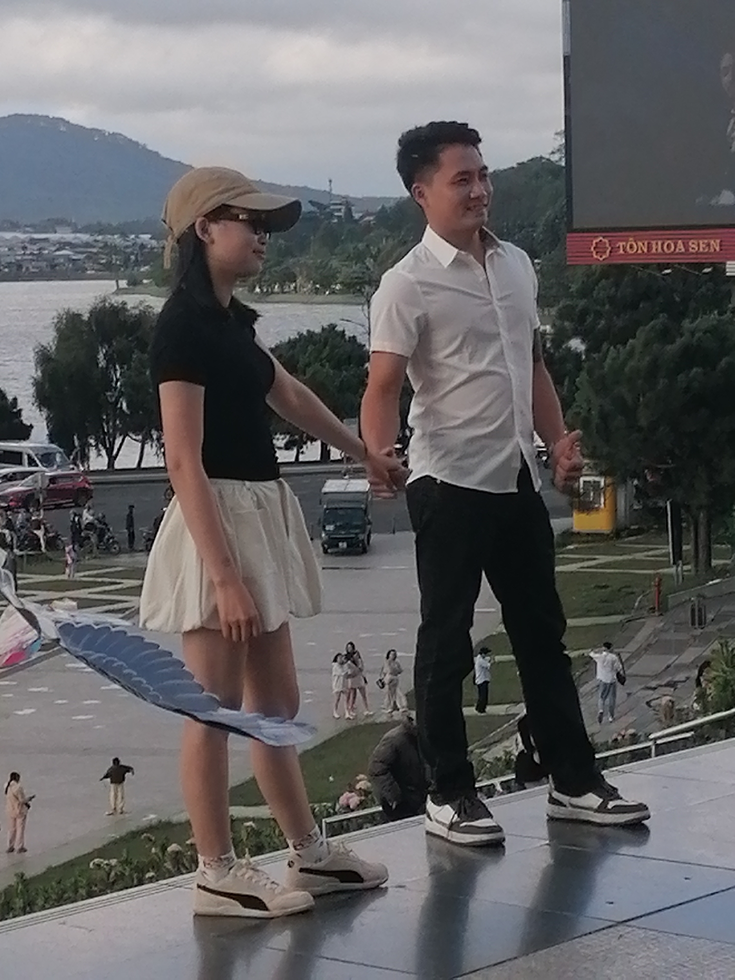 The image captures a couple holding hands and standing on a raised platform overlooking a body of water and a city. The woman, on the left, wears a black polo shirt, a cream-colored skirt, a tan baseball cap, and glasses. She also has on white Puma sneakers with black accents and patterned socks. The man, on the right, is dressed in a white button-down shirt and black pants, complemented by black and white sneakers. They are positioned in the foreground, with the expansive landscape of Da Lat, Vietnam, stretching out behind them. The background reveals a city nestled amongst hills, a tranquil lake, and lush green trees. On the left side of the image, a large, stylized kite, possibly resembling a fish, is partially visible. In the midground, a road with vehicles and people can be observed, along with a prominent billboard displaying Vietnamese text that reads "TÔN HOA SEN." The overall scene suggests a tourist spot or scenic overlook in Da Lat, likely during the day with overcast skies.