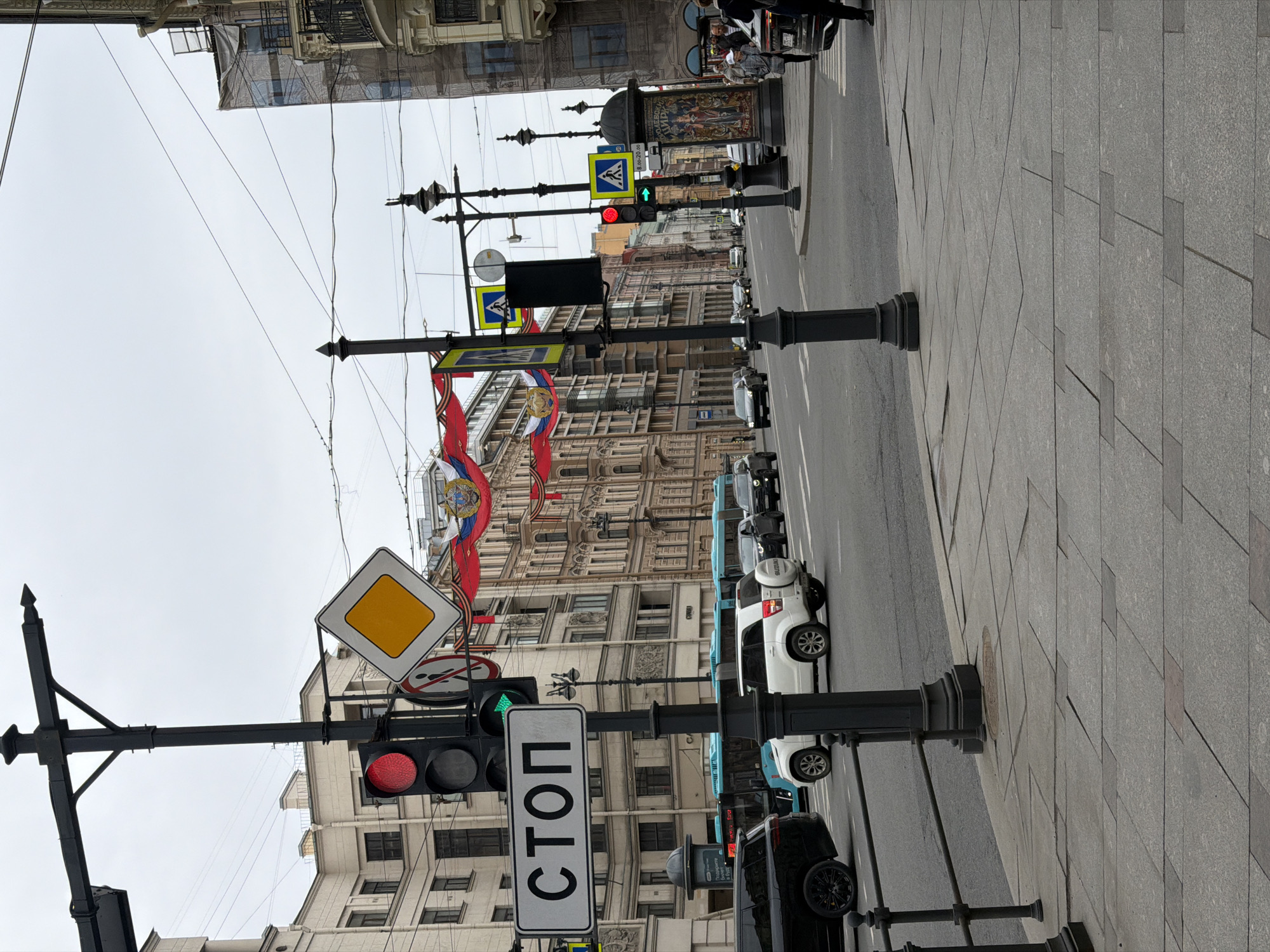 The image presents an urban street scene in Saint Petersburg, Russia, under an overcast sky during daytime. The view is taken from a sidewalk, looking down a city street lined with historical buildings.
In the foreground, several dark metal poles support an array of traffic infrastructure. A prominent red traffic light for vehicles is visible, below which hangs a large rectangular white sign with the Cyrillic word "СТОП" (STOP). Above, a yellow diamond-shaped sign indicates the main road priority. Further down the street, another traffic light displays a green signal for pedestrians. Other visible signs include blue square pedestrian crossing signs and a round "no entry" sign.
The street is flanked by grand, multi-story buildings exhibiting ornate facades and numerous windows, characteristic of Saint Petersburg's architectural style. Some buildings have scaffolding covered in white sheeting, indicating ongoing renovation or construction. Festive red banners, decorated with gold crests and St. George ribbons, are strung across the street, suggesting a celebratory or commemorative event, possibly Victory Day.
The street accommodates various vehicles, including a white Suzuki Jimny SUV, a black SUV, and several light blue vans or buses in the distance. A few pedestrians are visible on the sidewalk in the background, near a parked car and a cylindrical advertising pillar displaying an illegible poster. Overhead, a dense network of power lines spans across the street. A sign showing "8-20" is visible further down, possibly indicating hours of operation or a time restriction. The sidewalk itself is paved with large grey tiles.