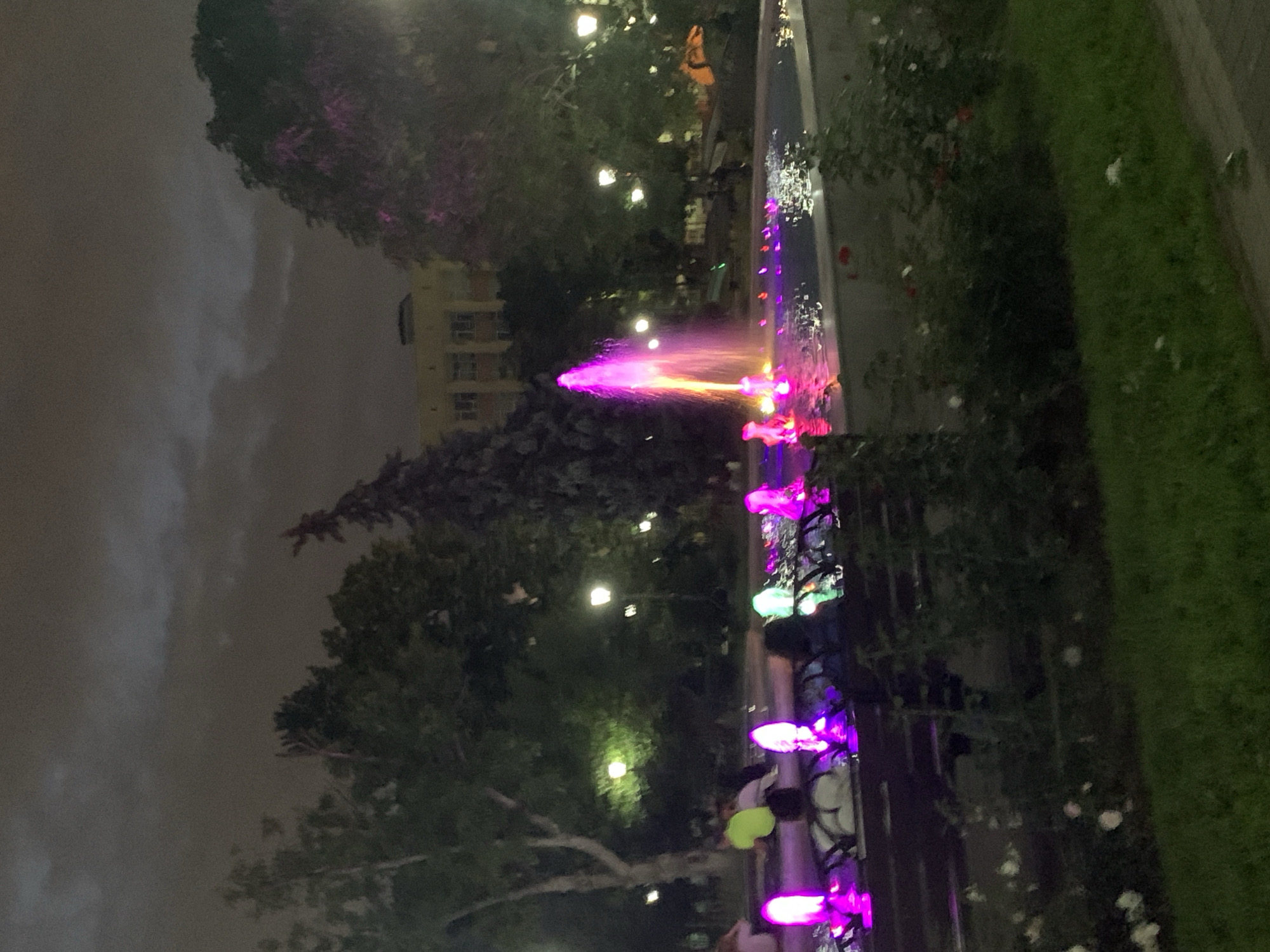 This is a nighttime outdoor scene of a park with a fountain. The fountain is illuminated with vibrant pink and purple lights, creating a colorful display in the water. Several people are sitting on benches around the fountain, some silhouetted against the lights. Trees surround the park, and a building with illuminated windows is visible in the background. The sky is dark and cloudy.