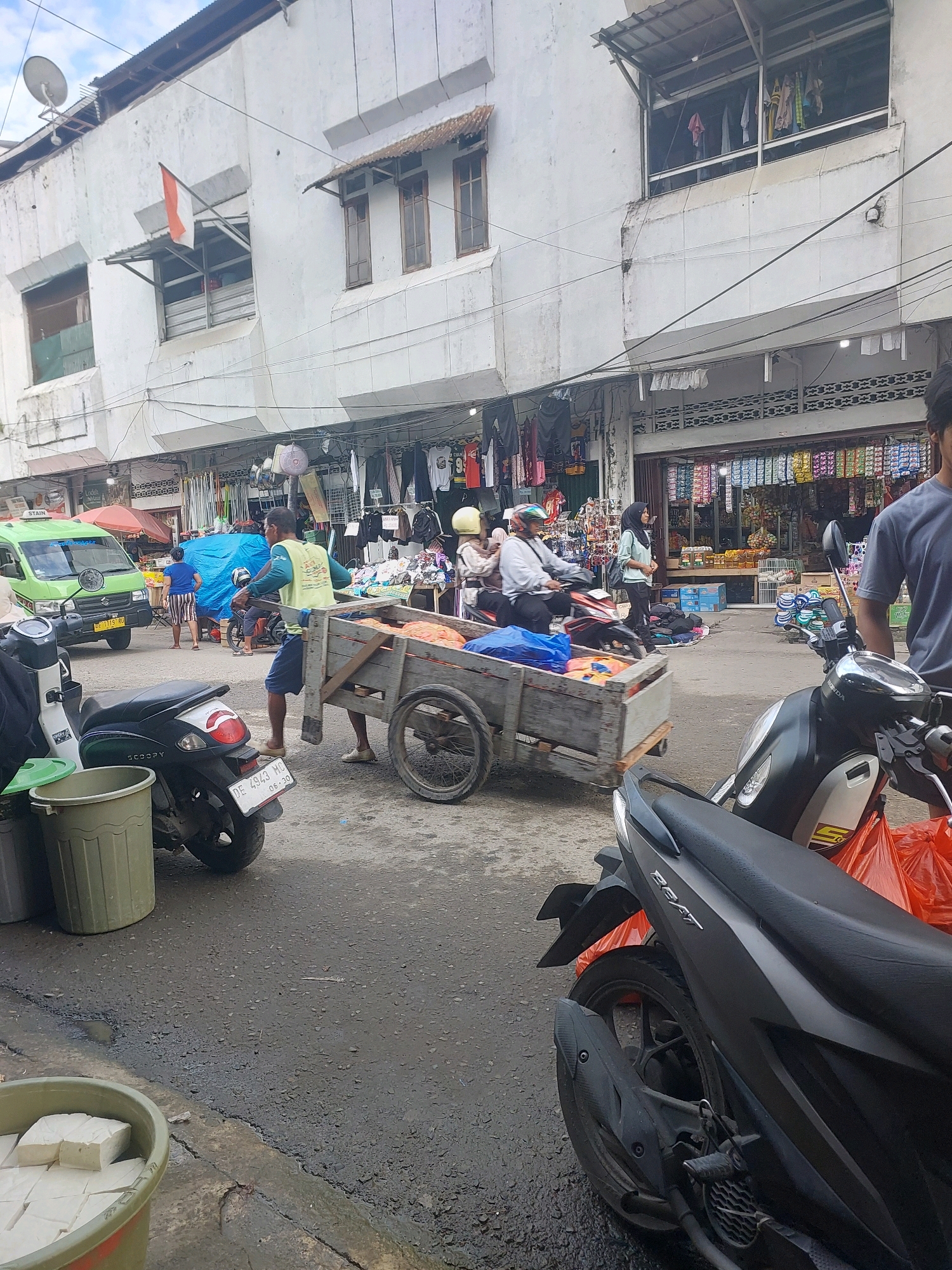 The image captures a street scene in Ambon, Indonesia, likely a market area given the presence of stalls and goods. In the foreground, a black motorcycle is parked on the wet asphalt, with an orange plastic bag behind it. To the left, a green trash bin with a lid is visible, and in the lower left corner, a green bin contains white blocks of what appears to be tofu.
A man in a yellow vest and blue shorts is pushing a wooden cart laden with orange material, possibly textiles or produce, along the street. Behind him, a green van is parked. Further back, a blue tarp covers a section of the street, and several people are visible, some on motorcycles. The buildings lining the street are two-story structures with visible awnings and clothing displayed for sale in some of the ground-level shops. The sky above is overcast.