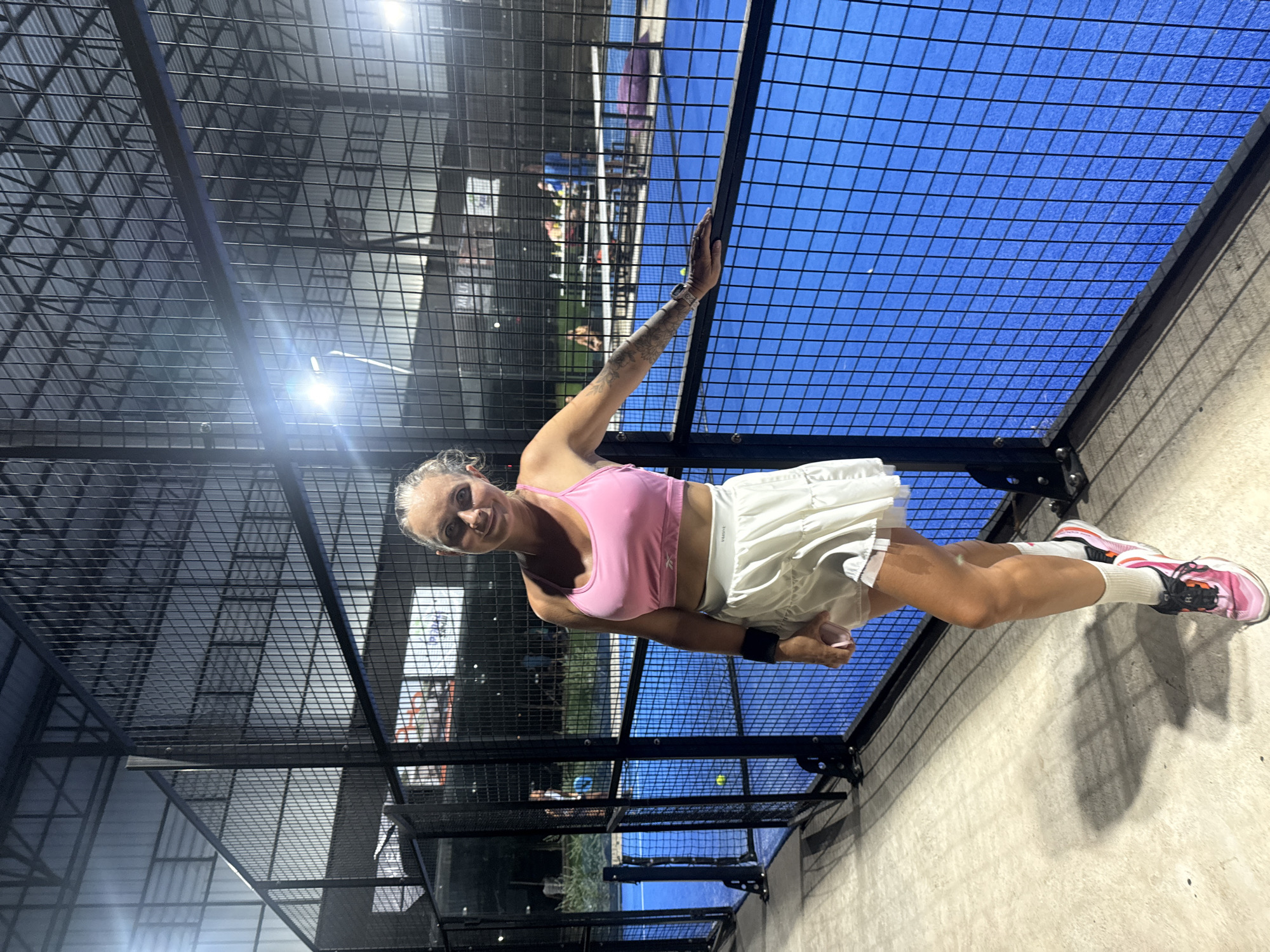 A woman is posing for a photo on the side of a padel court at night. The court is illuminated by artificial lights. The woman is wearing a pink sports bra, a white skirt, and athletic shoes. She is leaning against the fence surrounding the court, with her right arm resting on it. Her left hand is holding a phone. In the background, other people can be seen playing on the court or sitting at tables. The environment appears to be an outdoor sports facility with a covered roof structure. The image is taken from a low angle, focusing on the woman and the court.
