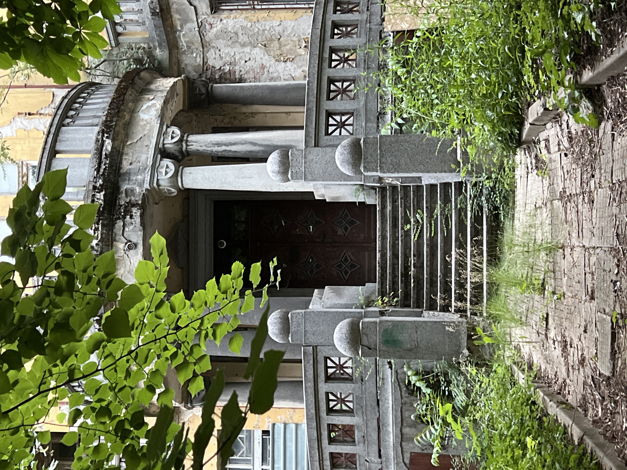 The image captures the entrance to an abandoned building, likely a former grand residence, located in Sofia, Bulgaria. The scene is overgrown with lush green foliage, with branches and leaves from trees in the foreground obscuring parts of the facade.
The building itself shows significant signs of decay. The facade is crumbling, with peeling paint revealing the underlying brickwork. A curved balcony, supported by classical columns with ornate capitals, extends from the upper floor. The railing of the balcony is decorated with a patterned design of squares containing star-like shapes.
A set of stone steps leads up to a large, dark wooden double door. The steps are flanked by stone pillars topped with spherical finials. The path leading to the steps is paved with worn bricks, cracked and uneven, with weeds and debris scattered across it. Abundant greenery, including tall grasses and leafy plants, surrounds the steps and the entrance, further emphasizing the building's abandonment and the encroachment of nature. The overall impression is one of faded grandeur and neglect.