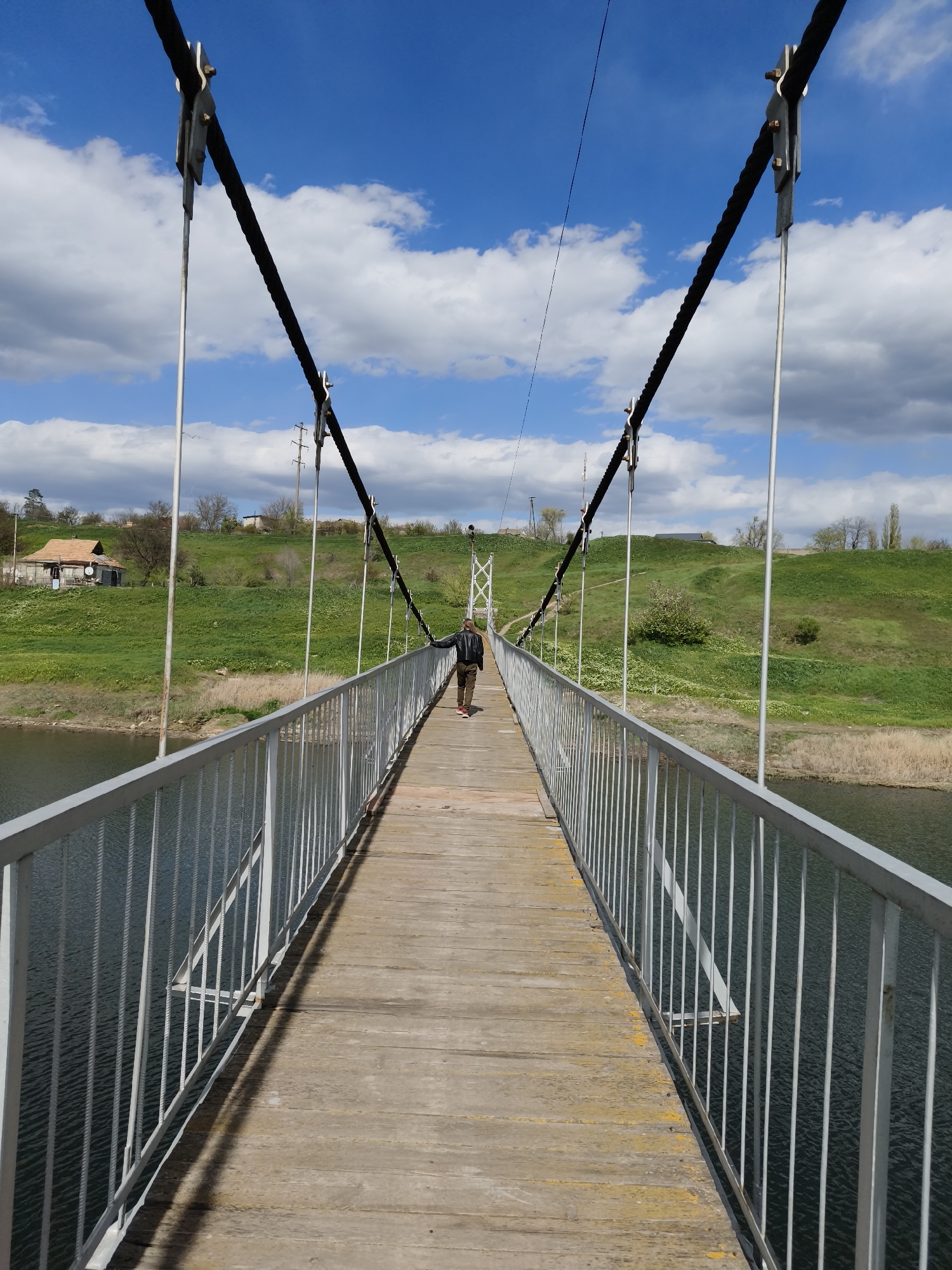 The media file captures a person walking on a suspension bridge over a body of water in a natural, semi-rural setting. The location is identified as Inhulets, Ukraine. The image is taken on a bright, sunny day, characterized by a clear blue sky dotted with fluffy white cumulus clouds.
The bridge itself has a weathered wooden deck and grey metal railings on either side. Thick, dark main suspension cables run above the bridge, from which thinner, vertical metal cables connect down to the deck structure, secured by metal plates and bolts. The water beneath the bridge is dark and shows gentle ripples.
On the left side of the frame, beyond the water, a lush green, grassy hill rises, featuring a small house with an orange-brown roof nestled among trees and bushes. The right side also presents verdant green hills with scattered foliage, and in the distance, what appears to be a power line structure can be seen.
A solitary figure, dressed in a dark jacket and dark pants, is walking away from the camera along the wooden deck of the bridge, positioned slightly to the right of the center. The sunlight casts distinct shadows from the railings onto the bridge's surface, suggesting it is late morning or early afternoon. The overall scene conveys a sense of peacefulness and calm. There is no visible text in the image.
