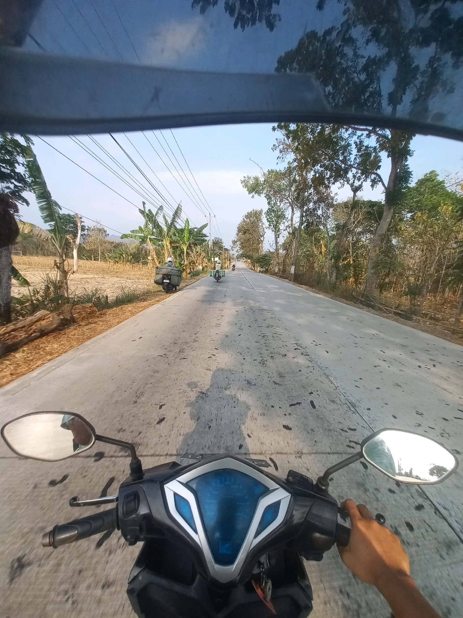 The image is a first-person perspective of someone riding a scooter on a paved road in Baturetno, Indonesia. The road is bordered by dry vegetation and trees on the right, and agricultural fields and banana trees on the left. In the distance, other motorbikes can be seen. Overhead, power lines stretch across the sky. The scooter's handlebars and instrument panel are visible in the foreground, with a hand gripping the right handlebar. Reflections of the sky and trees are visible in the rearview mirror. The overall environment suggests a rural or semi-rural setting under a clear sky.