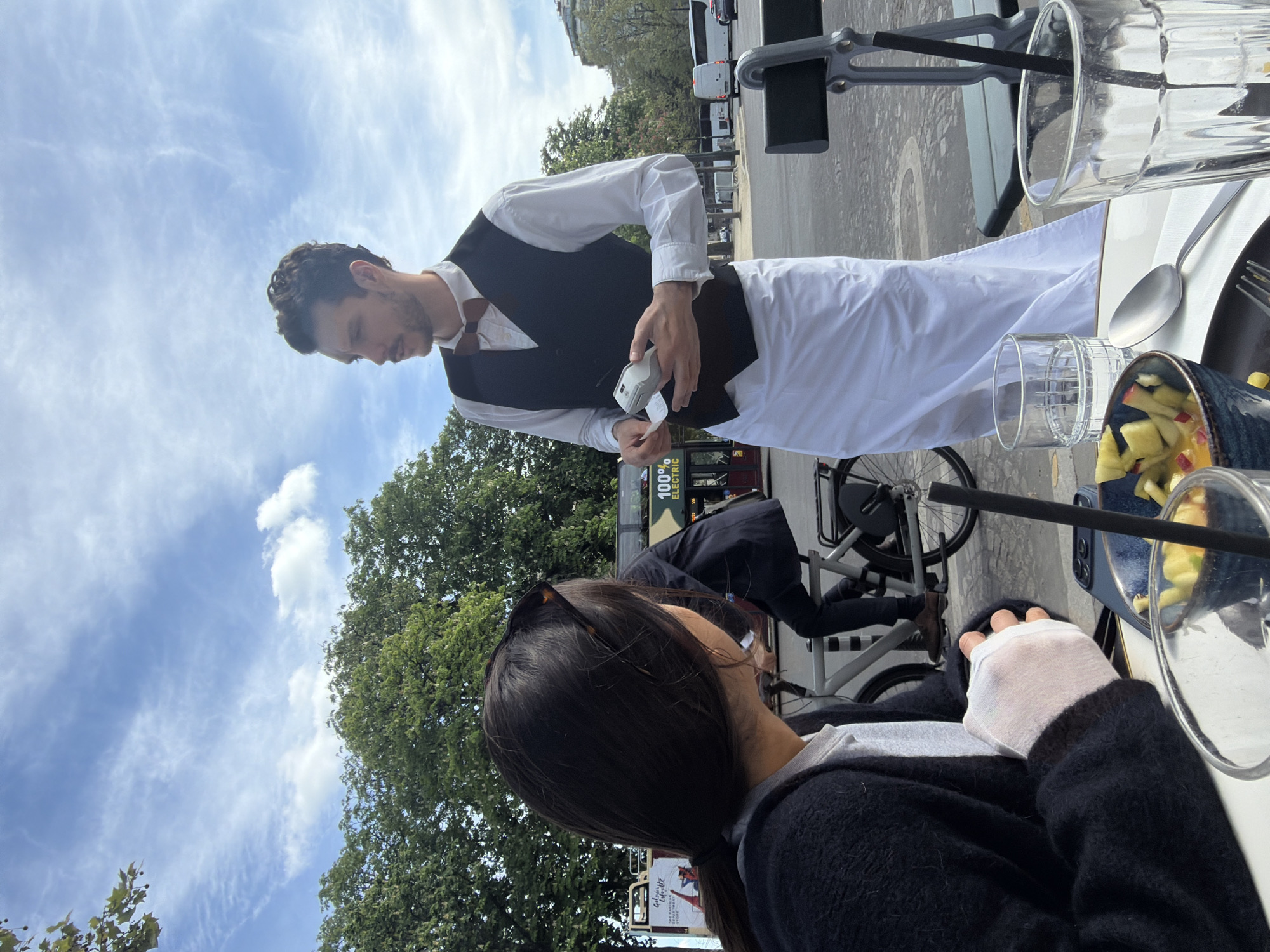 A server dressed in a black vest and white shirt is presenting a bill to a seated customer at an outdoor cafe. The customer has dark hair and is wearing a black sweater. A glass of water and a dish with diced fruit are on the table. A bicycle is parked nearby. The location appears to be a city street with trees and a bus in the background.