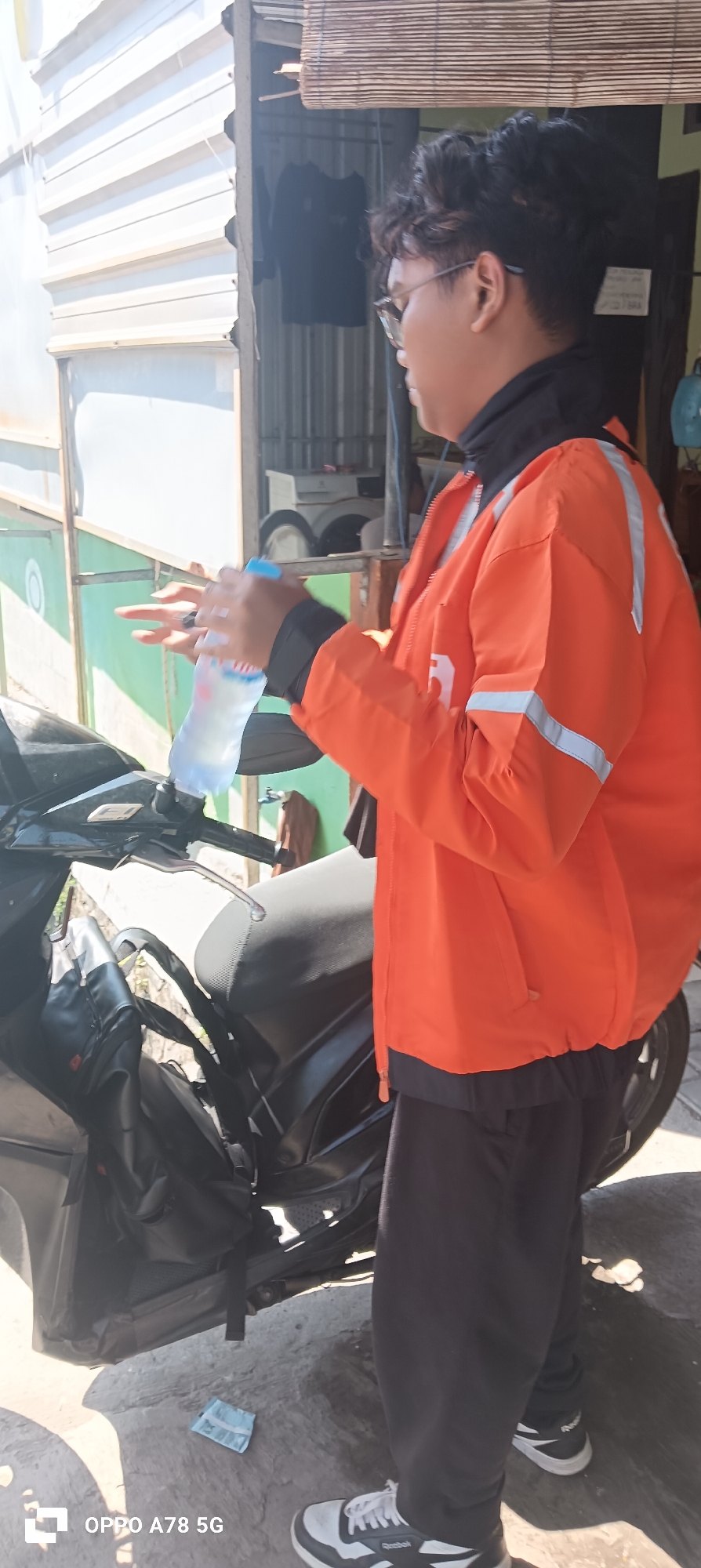 A person wearing an orange jacket with reflective stripes and black pants is standing next to a black motorcycle in what appears to be a residential area in Buncitan, Indonesia. The person is holding a bottle of water and appears to be in the process of drinking from it. The motorcycle has a black bag attached to the back. The background shows a building with corrugated metal siding and a bamboo-slatted window covering. The ground is paved with concrete and there are some loose papers on the ground. The lighting suggests it is daytime, likely sunny, casting shadows. The person has dark hair and is wearing glasses.